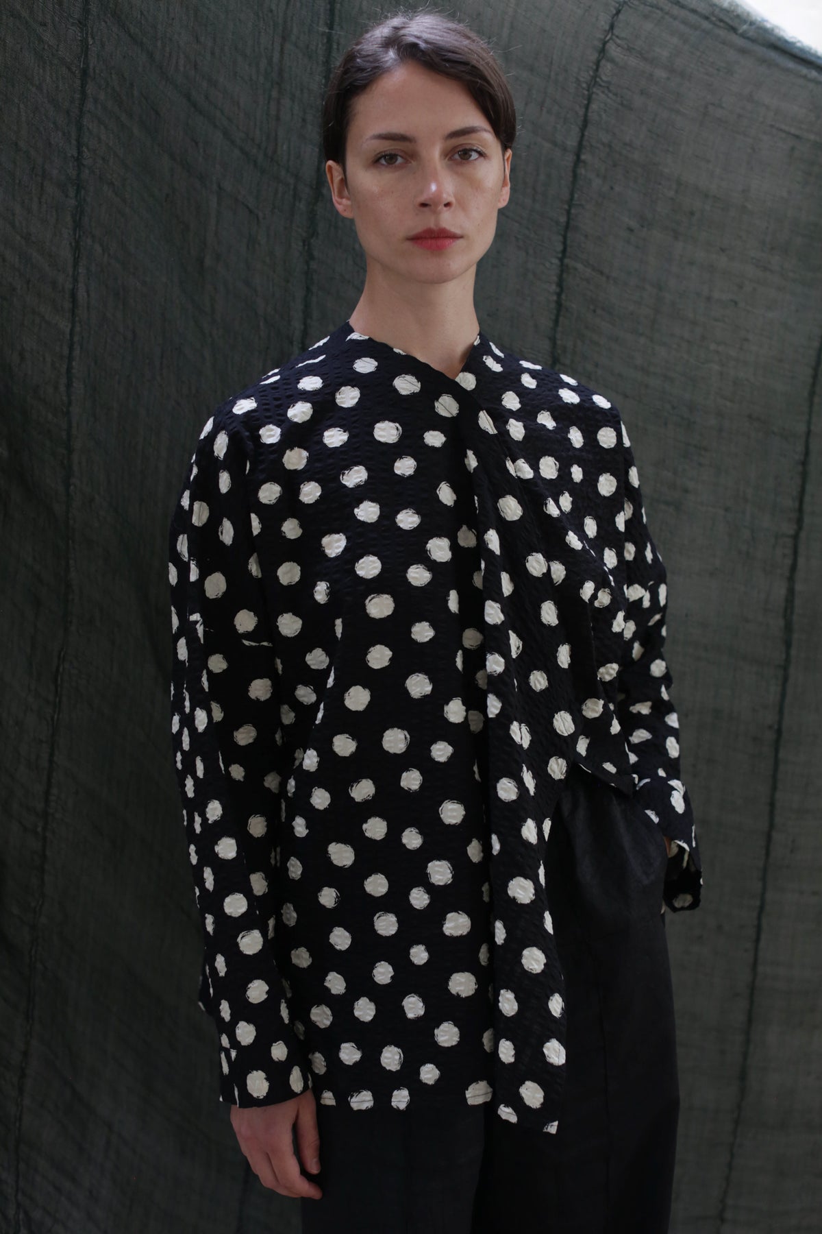 A person with dark hair pulled back wears the Qipao Blouse | Polkadot Seersucker Black, paired with black pants, standing before a dark textured background.