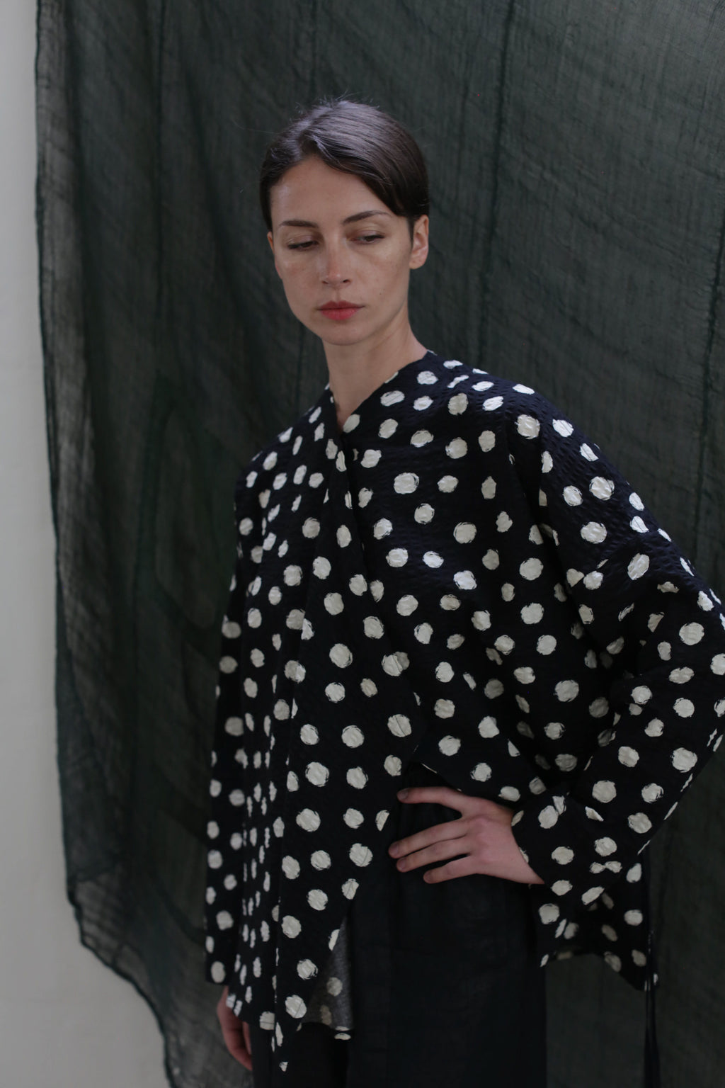 A woman with short dark hair models the Qipao Blouse | Polkadot Seersucker Black, standing in front of a dark green textured backdrop with one hand on her hip, gazing slightly down to the side.