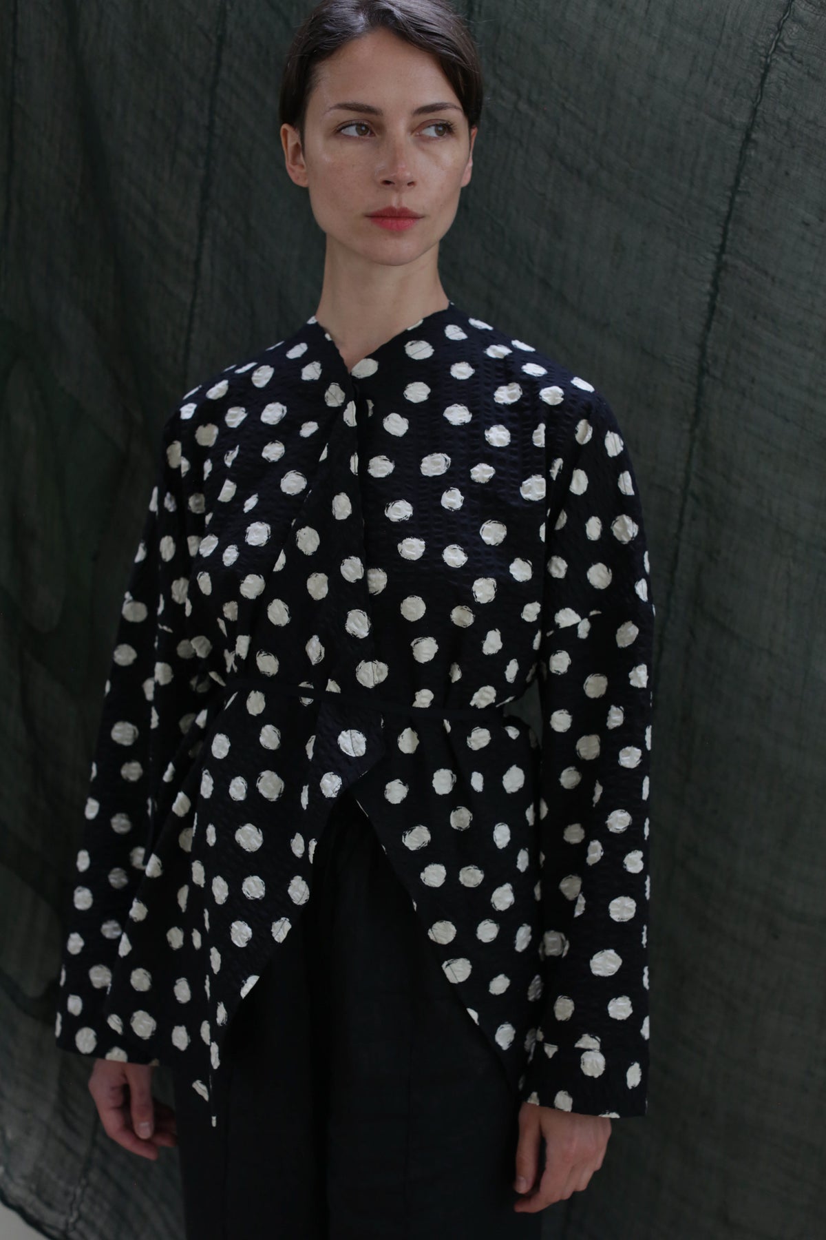 A woman stands before a dark, textured background, wearing the Qipao Blouse | Polkadot Seersucker Black and black pants. She gazes slightly to the side with a neutral expression.