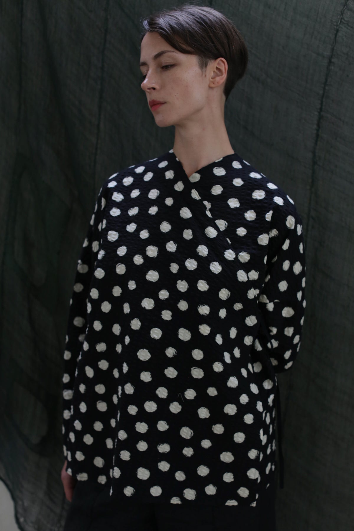A person with short brown hair stands before a dark green backdrop, wearing the Qipao Blouse | Polkadot Seersucker Black and looking down to the side with a neutral expression.
