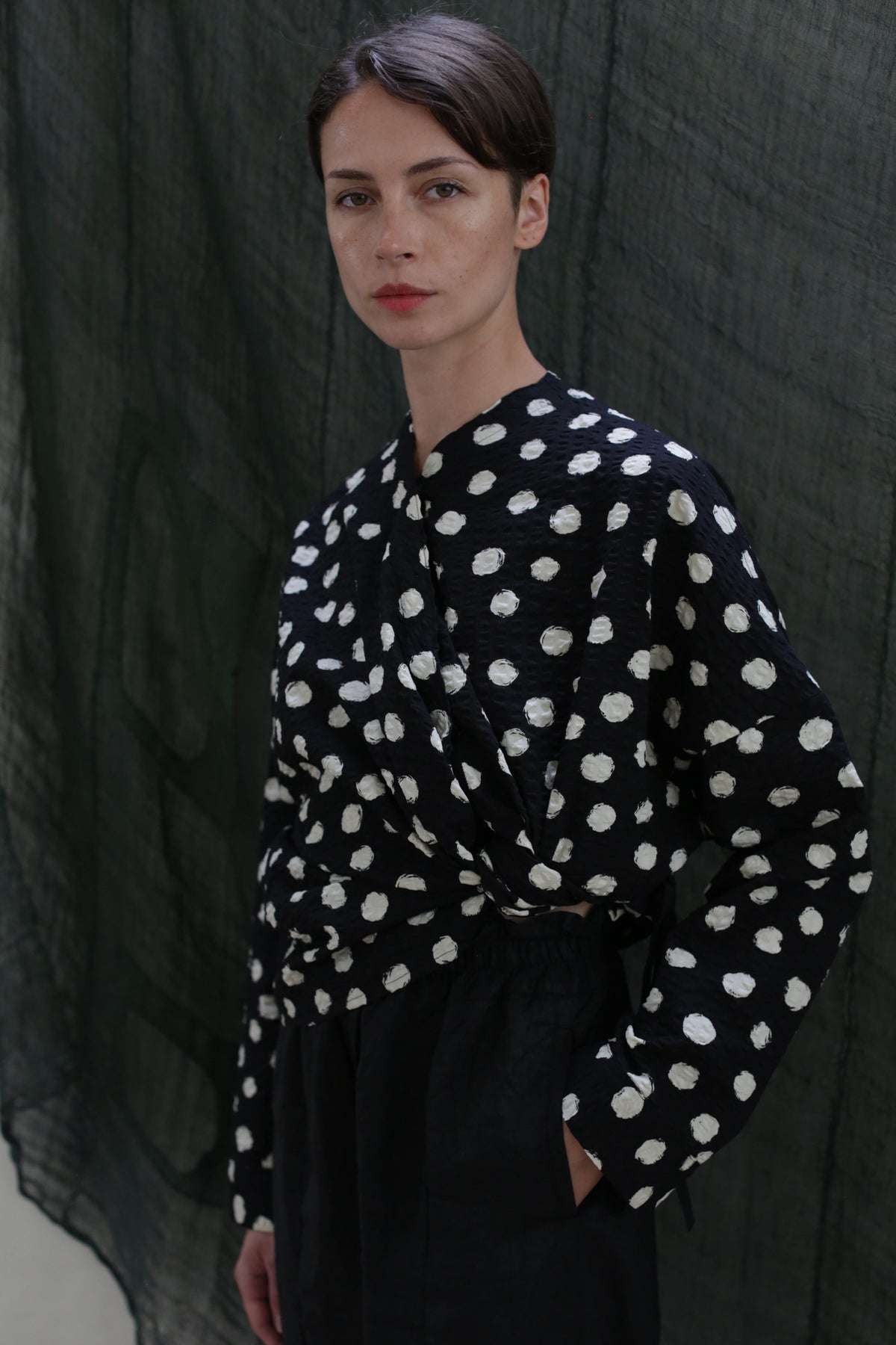 A woman with short brown hair stands before a dark green textured backdrop, calmly looking at the camera in the Qipao Blouse | Polkadot Seersucker Black and black high-waisted pants, one hand casually in her pocket.