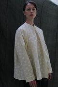 Against a dark fabric backdrop, a person with short hair and a neutral expression wears the Qipao Blouse | Polkadot Seersucker Butter—an oversized off-white top with a subtle polkadot pattern and wide sleeves.