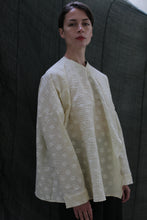 Load image into Gallery viewer, Against a dark fabric backdrop, a person with short hair and a neutral expression wears the Qipao Blouse | Polkadot Seersucker Butter—an oversized off-white top with a subtle polkadot pattern and wide sleeves.