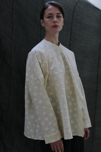 Against a dark fabric backdrop, a person with short hair and a neutral expression wears the Qipao Blouse | Polkadot Seersucker Butter—an oversized off-white top with a subtle polkadot pattern and wide sleeves.