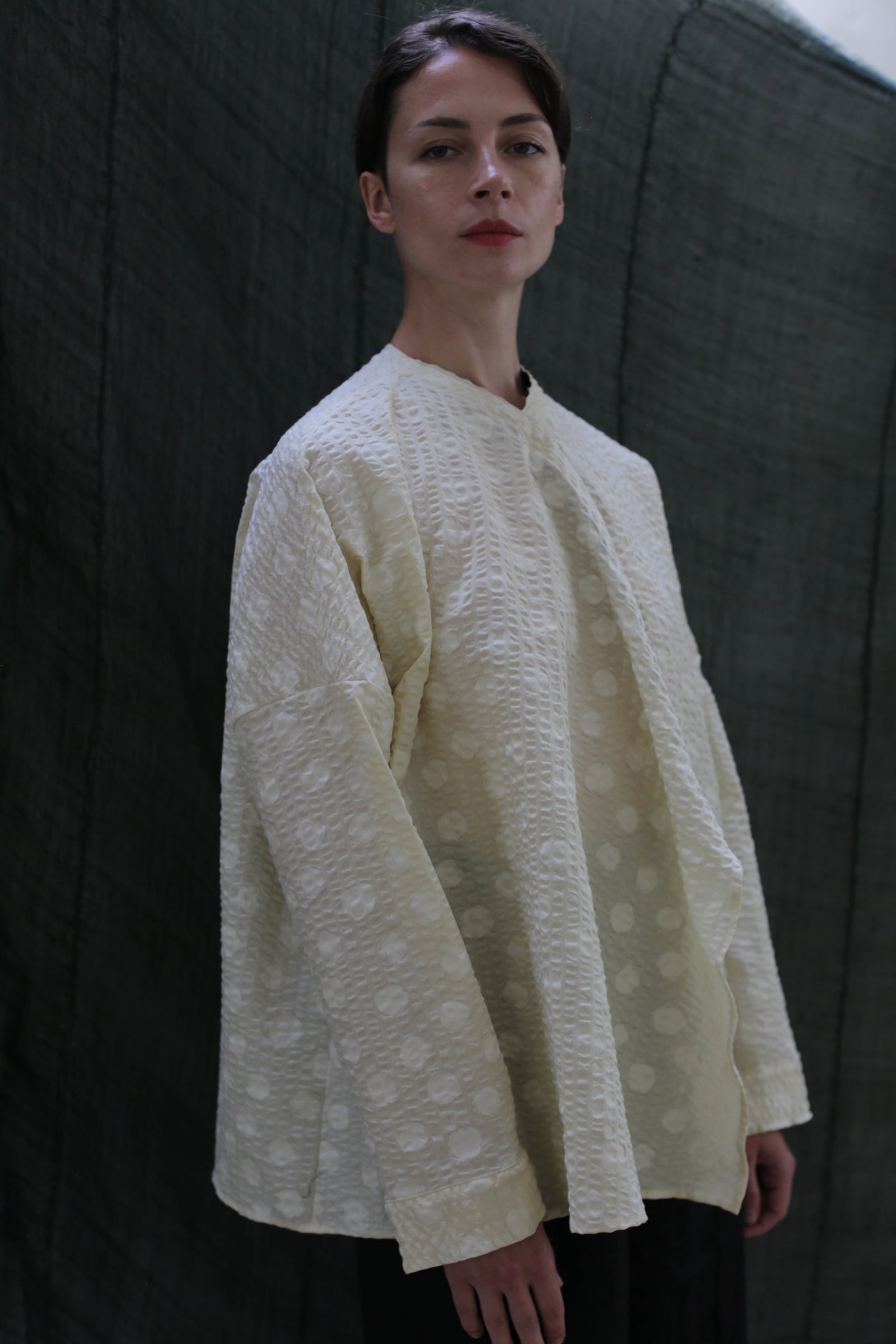 Against a dark fabric backdrop, a person with short hair and a neutral expression wears the Qipao Blouse | Polkadot Seersucker Butter—an oversized off-white top with a subtle polkadot pattern and wide sleeves.