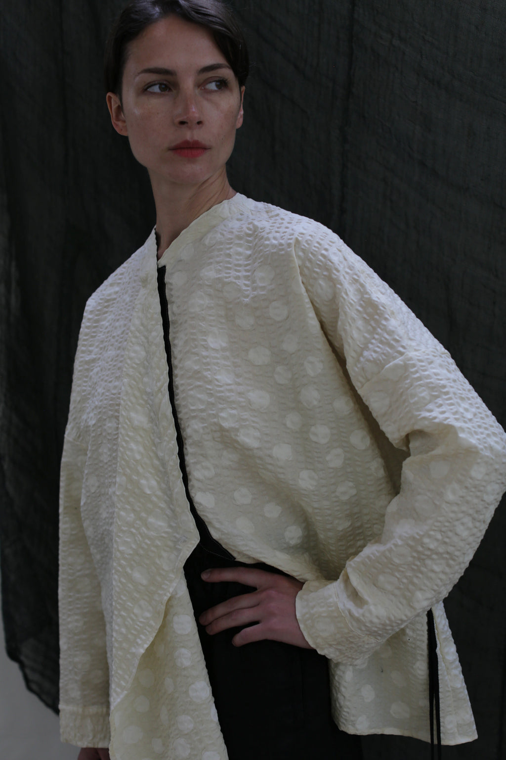 A person with short brown hair models the Qipao Blouse in Polkadot Seersucker Butter, featuring a textured cream fabric with subtle polka dots, standing against a dark draped background with one hand on their hip.