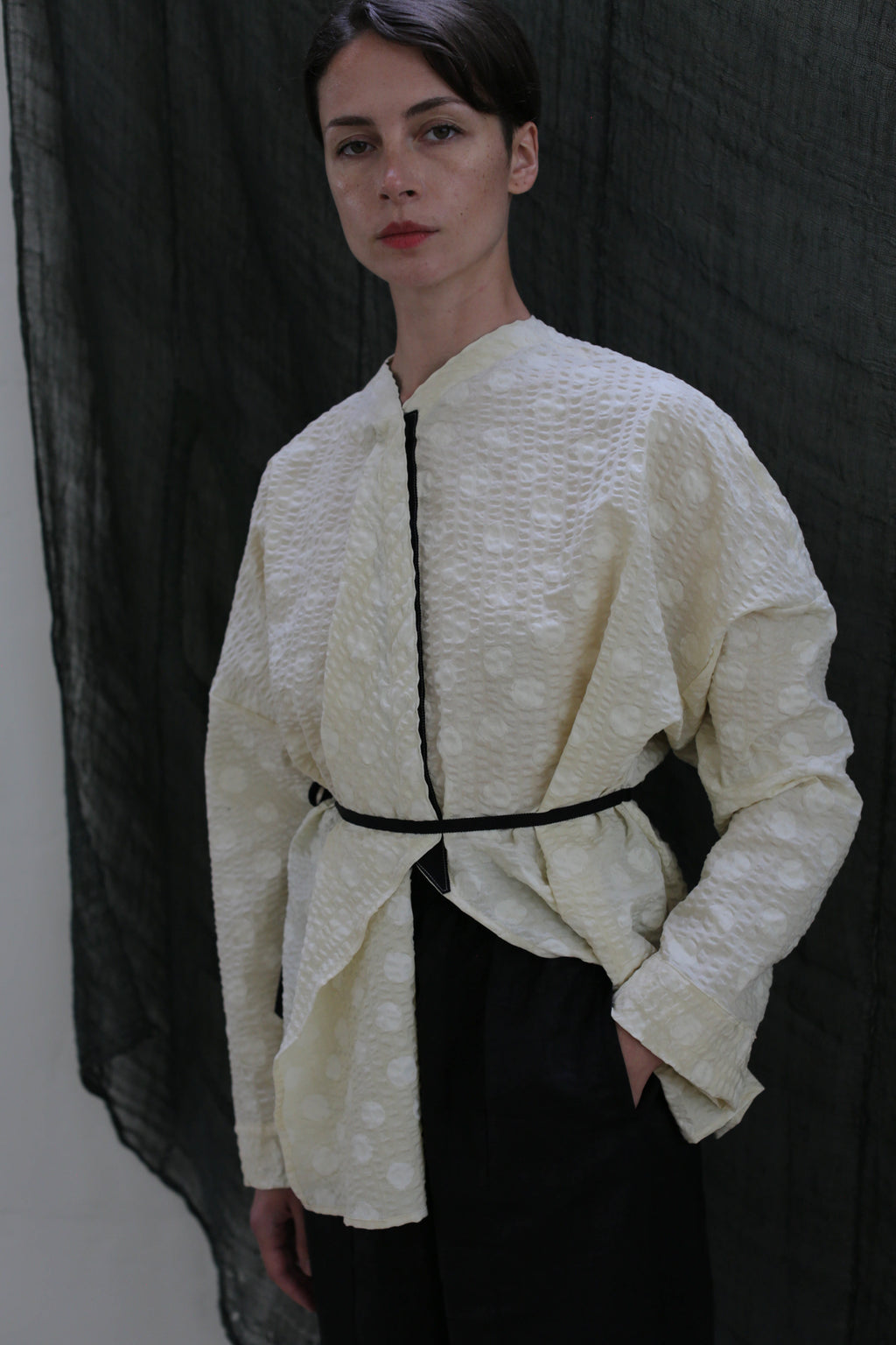 A person stands against a dark fabric backdrop, wearing the Qipao Blouse in Polkadot Seersucker Butter, styled oversized with a thin black belt at the waist and dark pants, one hand in their pocket.