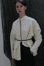 Load image into Gallery viewer, A person stands against a dark fabric backdrop, wearing the Qipao Blouse in Polkadot Seersucker Butter, styled oversized with a thin black belt at the waist and dark pants, one hand in their pocket.