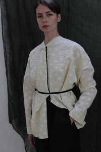 A person stands against a dark fabric backdrop, wearing the Qipao Blouse in Polkadot Seersucker Butter, styled oversized with a thin black belt at the waist and dark pants, one hand in their pocket.