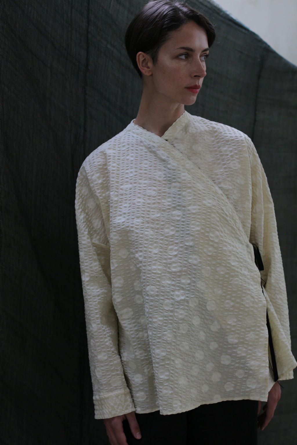 A person with short dark hair stands against a dark fabric backdrop, wearing the Qipao Blouse | Polkadot Seersucker Butter—a loose, cream-colored long-sleeve top with subtle polkadots—looking slightly to the right.