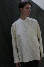 Load image into Gallery viewer, A person with short dark hair stands against a dark fabric backdrop, wearing the Qipao Blouse | Polkadot Seersucker Butter—a loose, cream-colored long-sleeve top with subtle polkadots—looking slightly to the right.