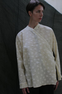 A person with short dark hair stands against a dark fabric backdrop, wearing the Qipao Blouse | Polkadot Seersucker Butter—a loose, cream-colored long-sleeve top with subtle polkadots—looking slightly to the right.