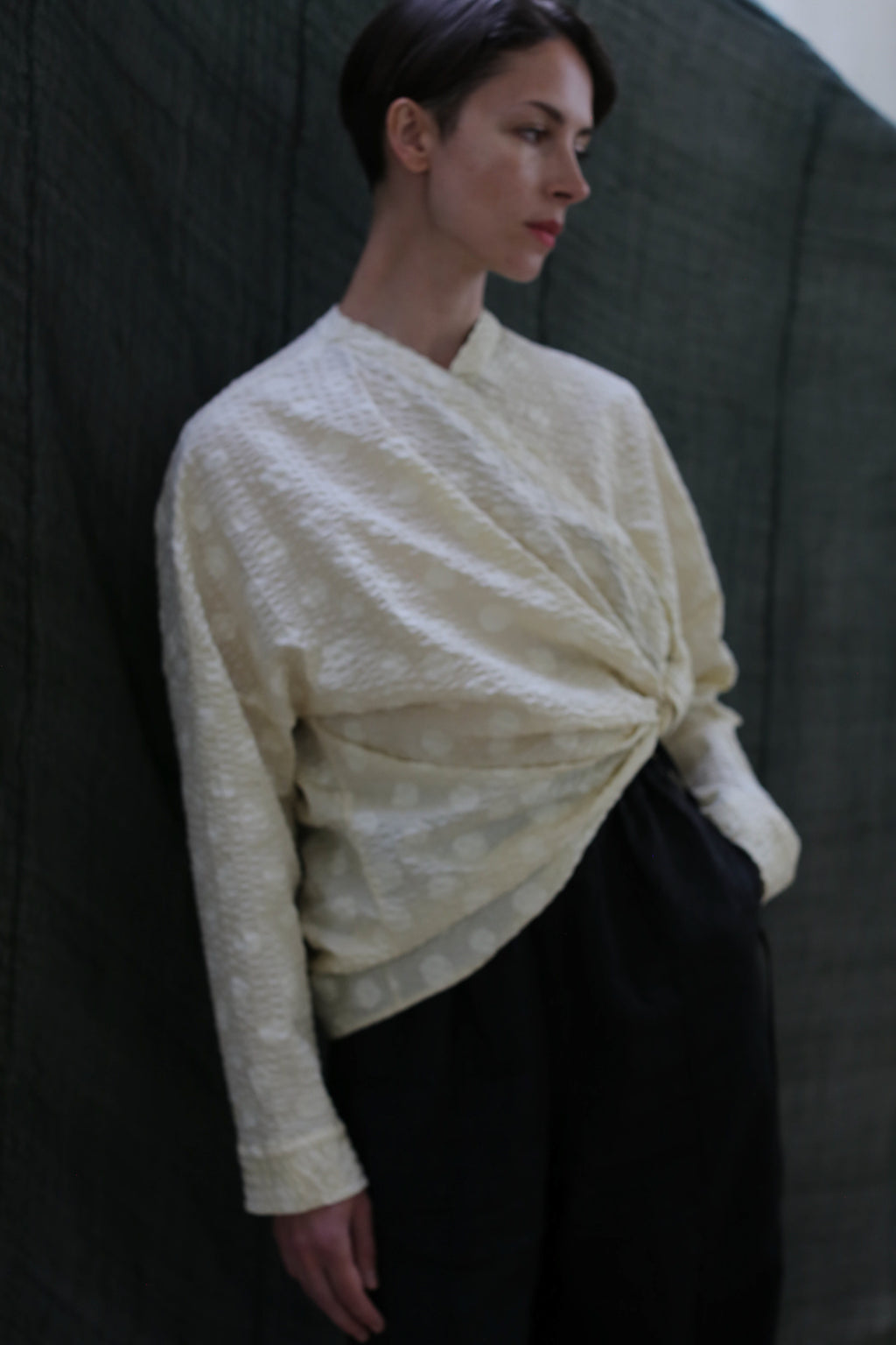 A person with short dark hair wears the Qipao Blouse | Polkadot Seersucker Butter and loose black pants, standing against a dark green fabric background.