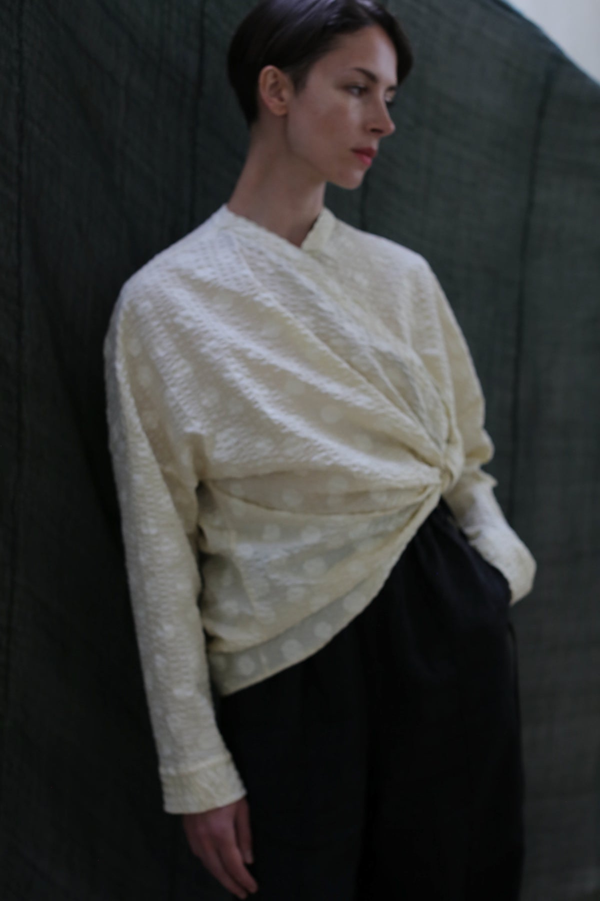 A person with short dark hair wears the Qipao Blouse | Polkadot Seersucker Butter and loose black pants, standing against a dark green fabric background.