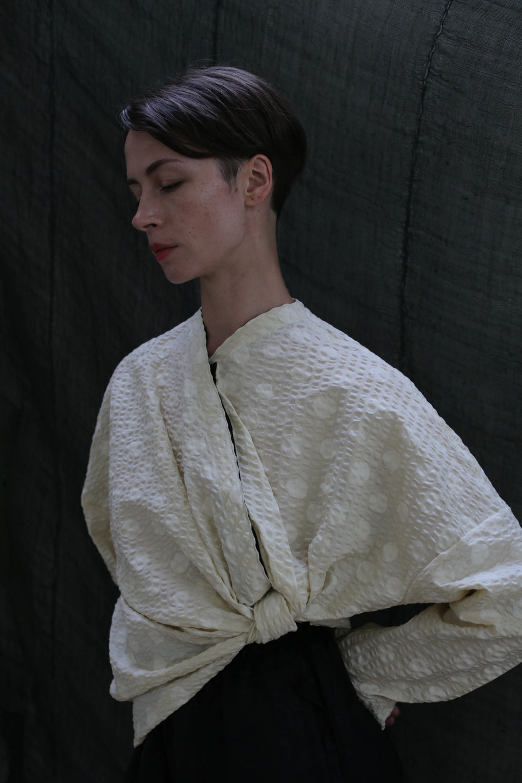 A person with short dark hair stands against a dark backdrop, eyes closed, wearing the Qipao Blouse in Polkadot Seersucker Butter, featuring wide sleeves tied in front.