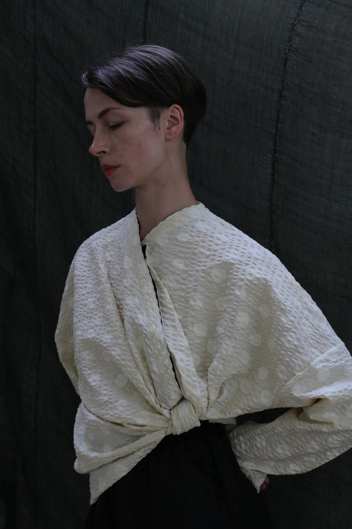 A person with short dark hair stands against a dark backdrop, eyes closed, wearing the Qipao Blouse in Polkadot Seersucker Butter, featuring wide sleeves tied in front.