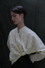 Load image into Gallery viewer, A person with short dark hair stands against a dark backdrop, eyes closed, wearing the Qipao Blouse in Polkadot Seersucker Butter, featuring wide sleeves tied in front.