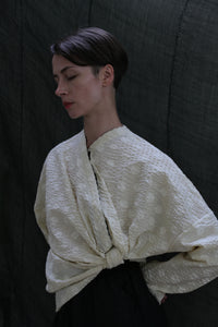 A person with short dark hair stands against a dark backdrop, eyes closed, wearing the Qipao Blouse in Polkadot Seersucker Butter, featuring wide sleeves tied in front.