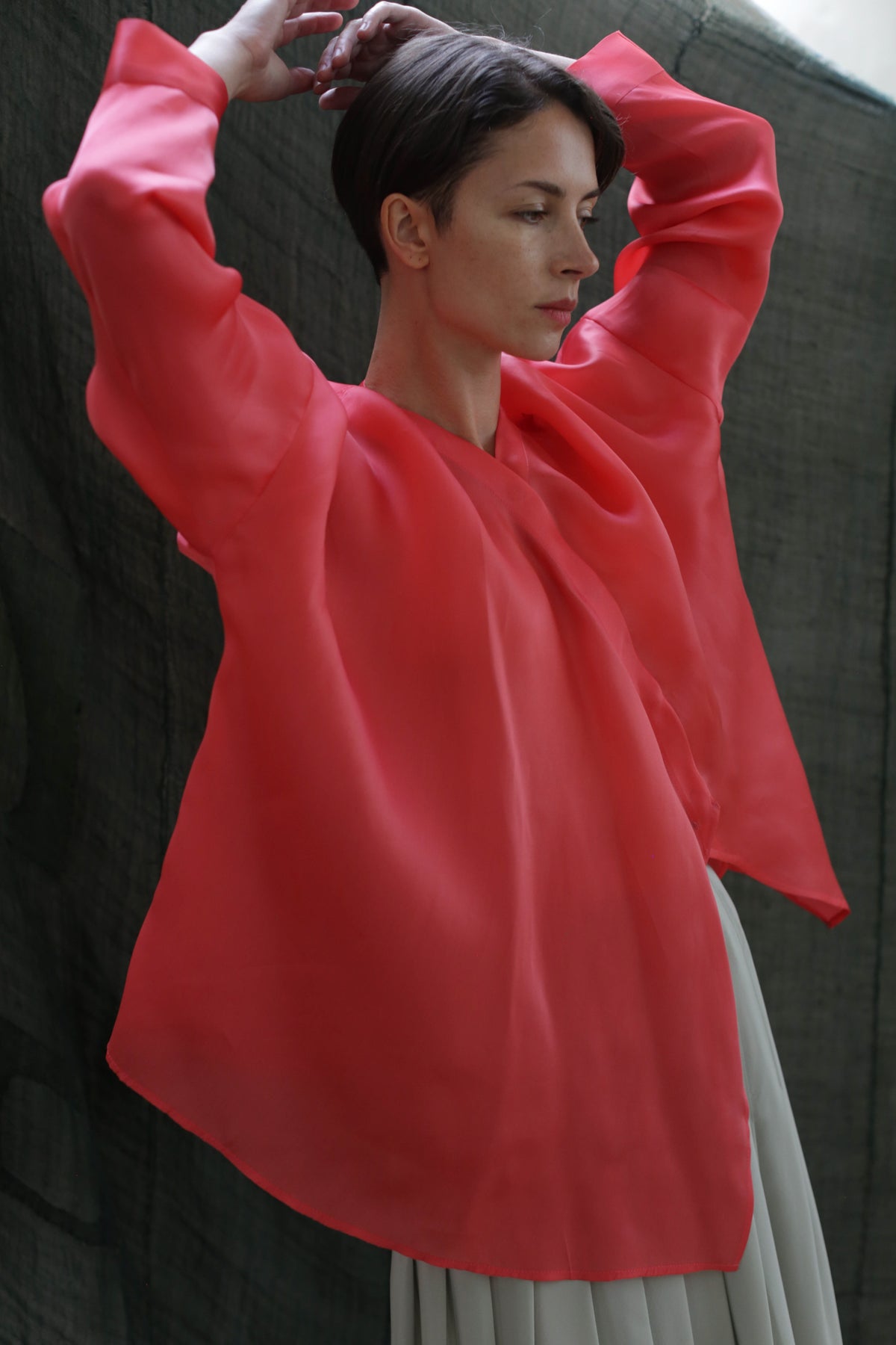 A person with short dark hair stands with arms raised, wearing the Qipao Blouse | Silk Organza Sakura in bright pink and light-colored pants, set against a dark, textured background.