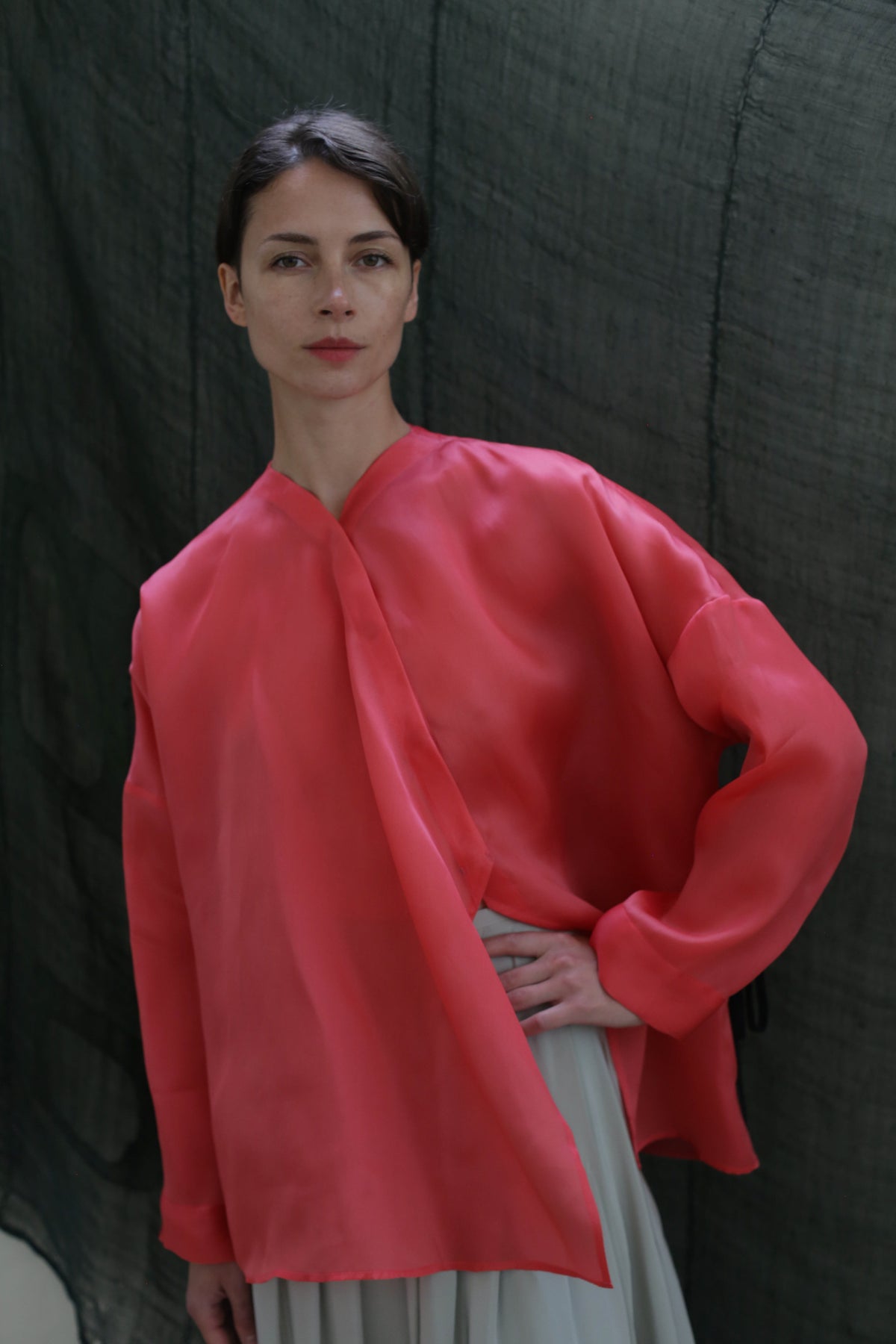 A woman poses with one hand on her hip, wearing the Qipao Blouse | Silk Organza Sakura in bright pink and a light skirt, set against a dark, textured background.