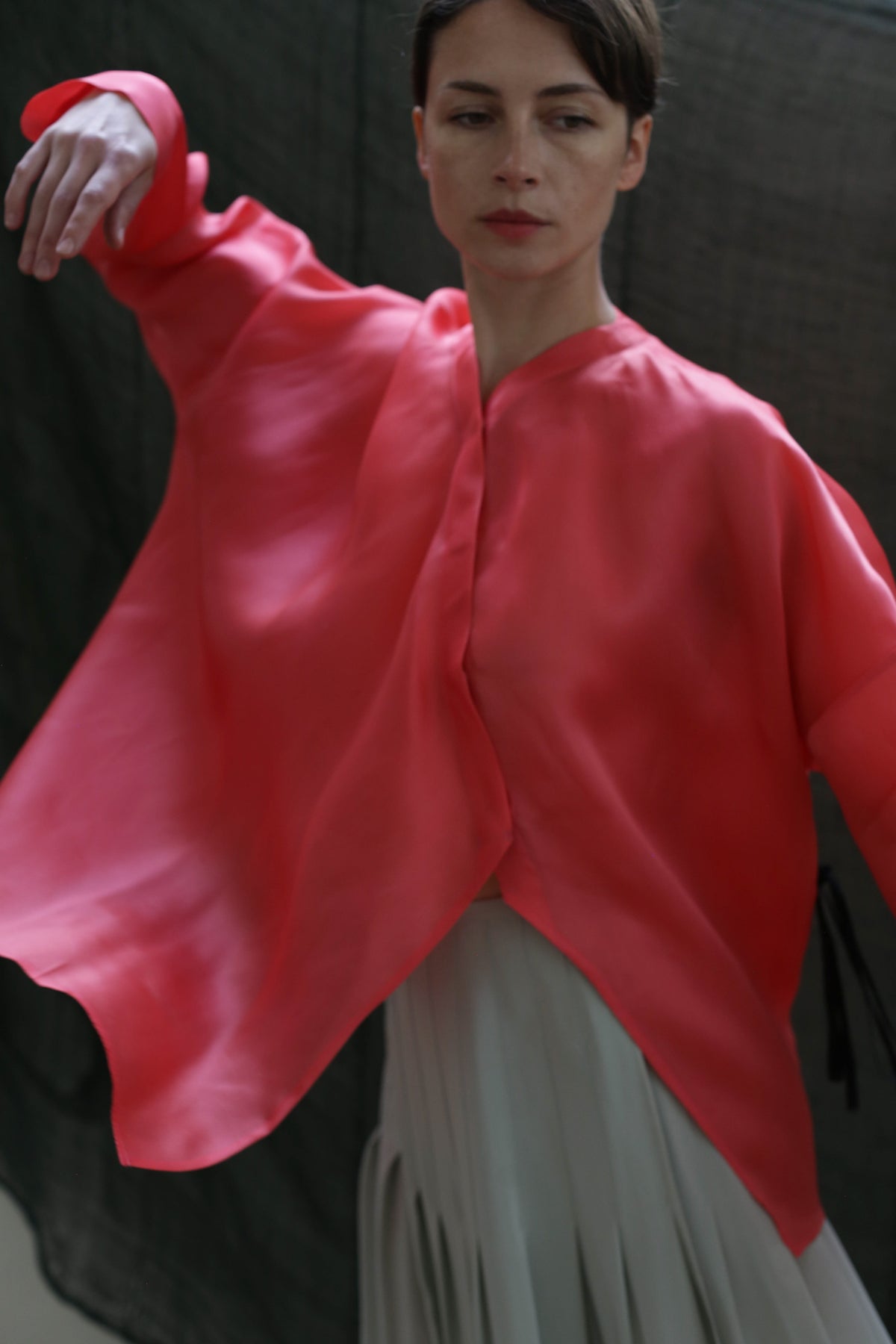 A person poses with one arm raised, wearing the flowing, bright pink Qipao Blouse | Silk Organza Sakura by Atelier Lan Jaenicke over a light skirt. The dark, blurred background highlights the fabric's elegant movement.