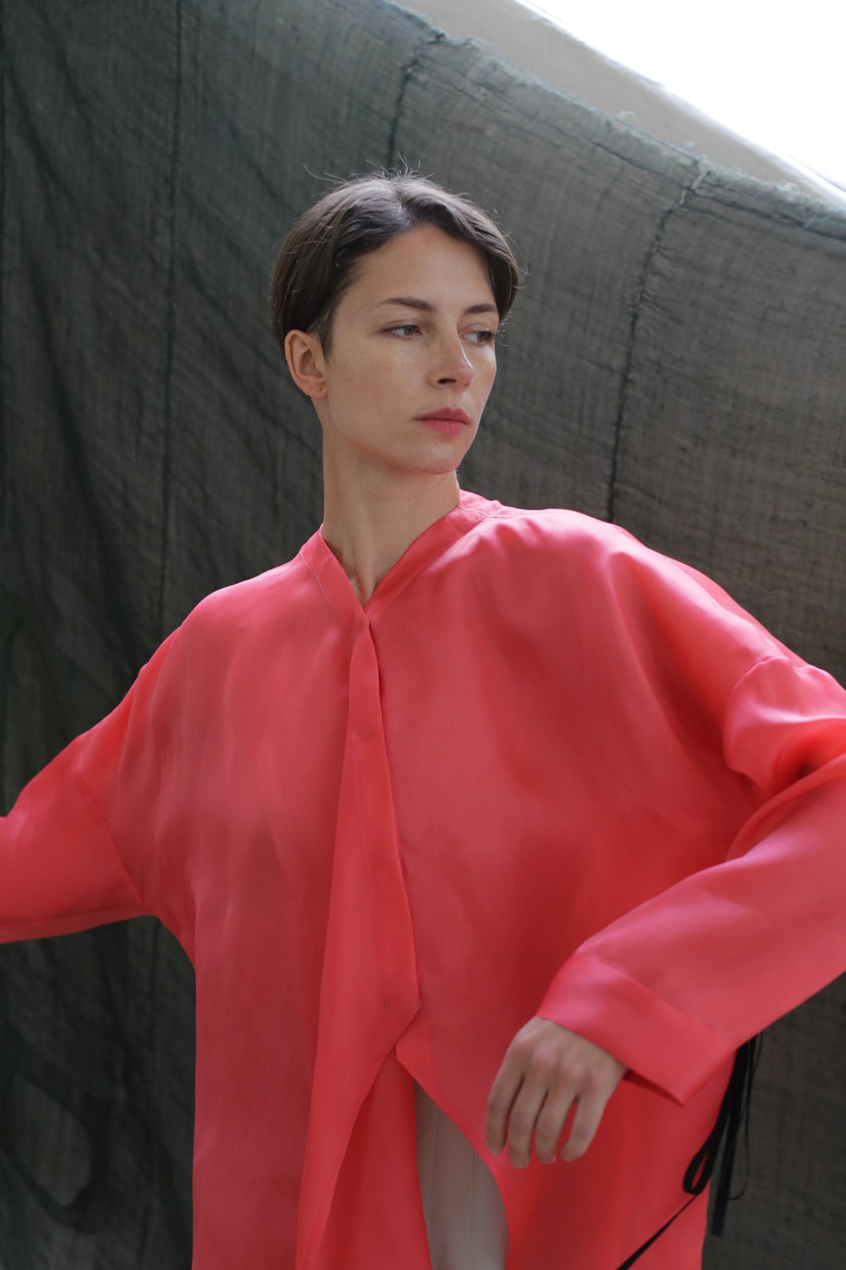 A person with short brown hair wearing a loose, bright pink Qipao Blouse | Silk Organza Sakura stands before a textured dark green backdrop, gazing to the side with a neutral expression.