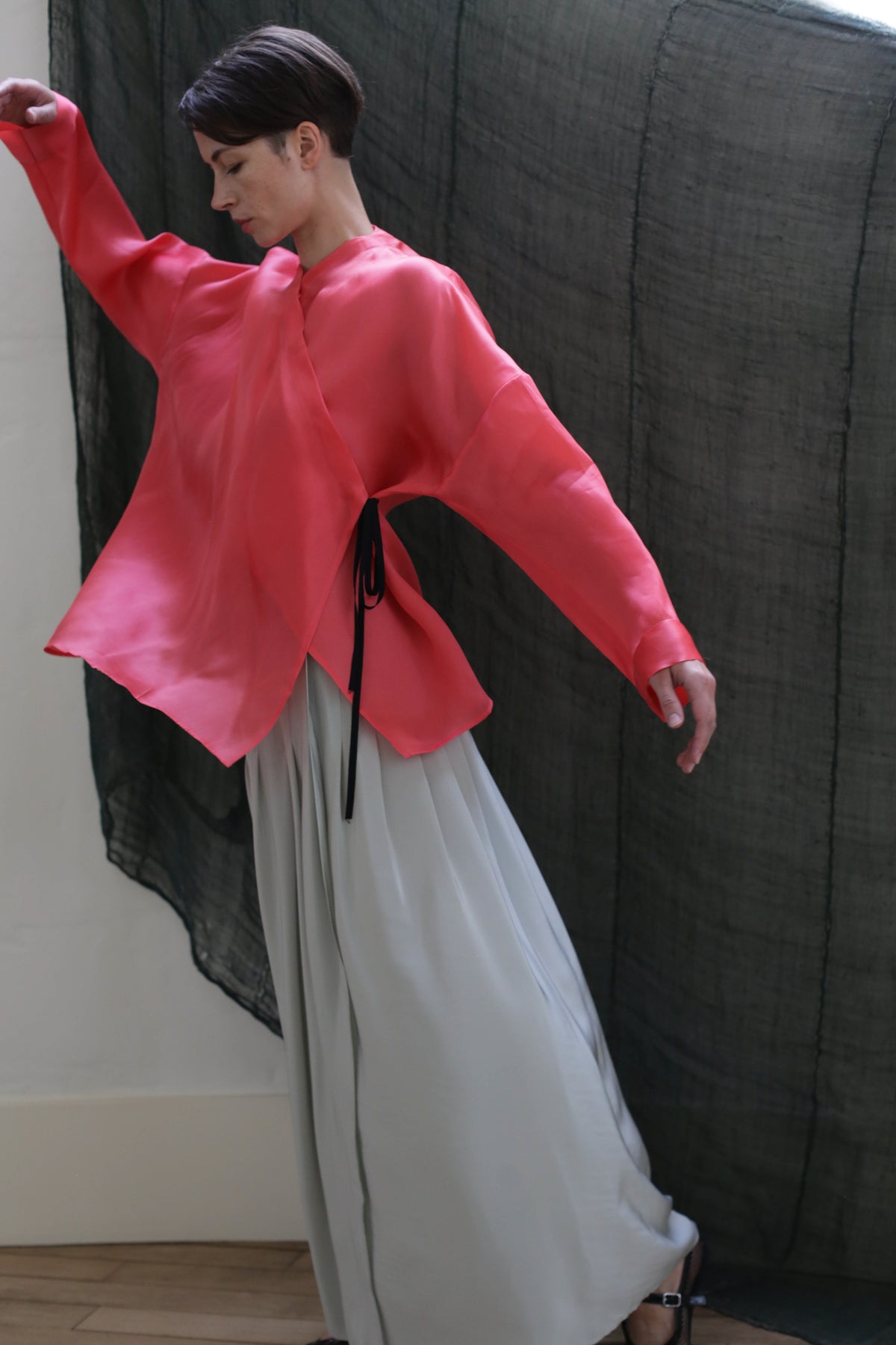 A person poses gracefully indoors, wearing the Hand Pleated Column Wrap Skirt in Crepe de Chine Celadon, styled with a loose top, standing before a dark sheer fabric backdrop.