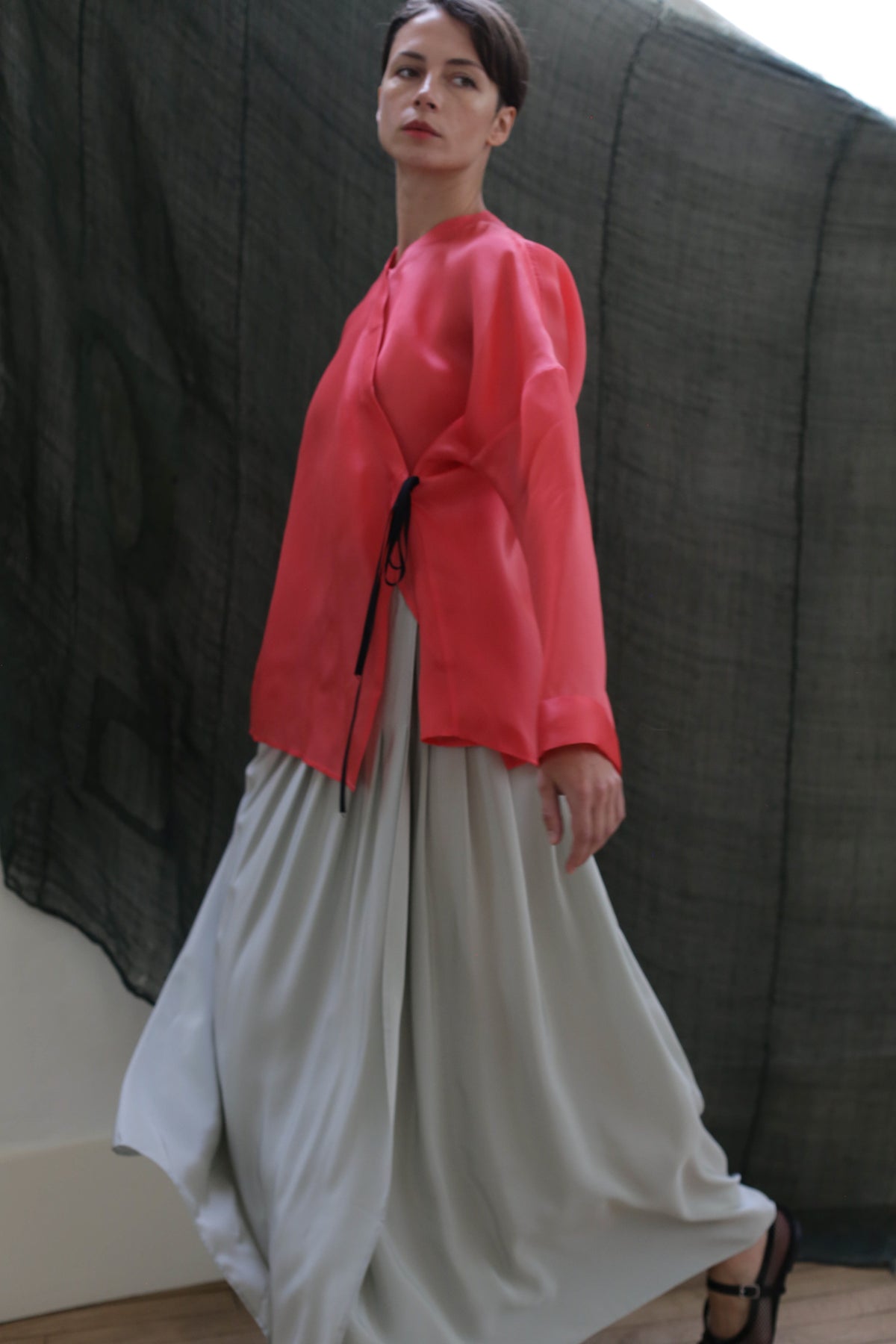 A woman in a loose, bright pink top and the Hand Pleated Column Wrap Skirt in Crepe de Chine Celadon walks indoors against a sheer black fabric backdrop.