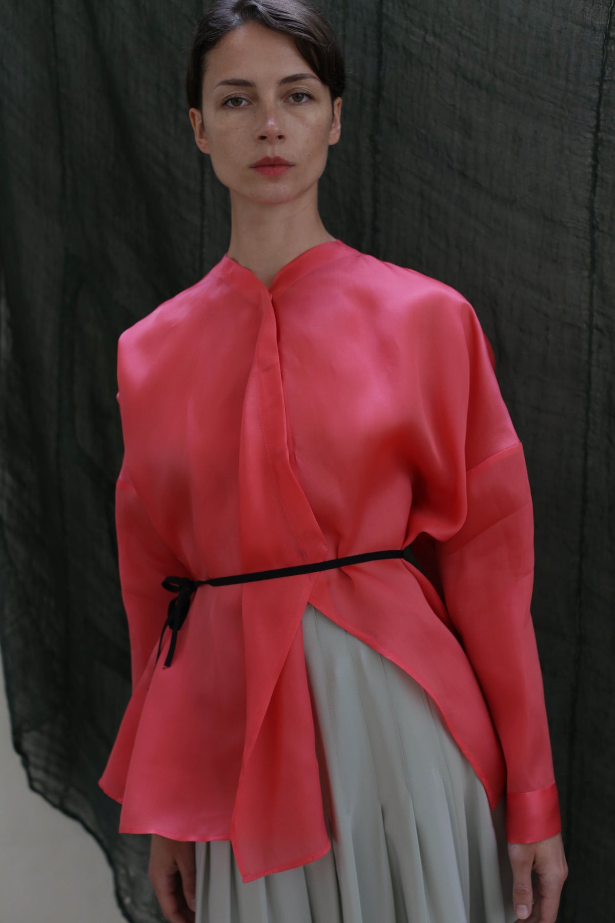 A woman with short hair and a neutral expression stands before a dark, textured backdrop, wearing the Qipao Blouse | Silk Organza Sakura—a loose, bright pink top belted at the waist with a thin black string.