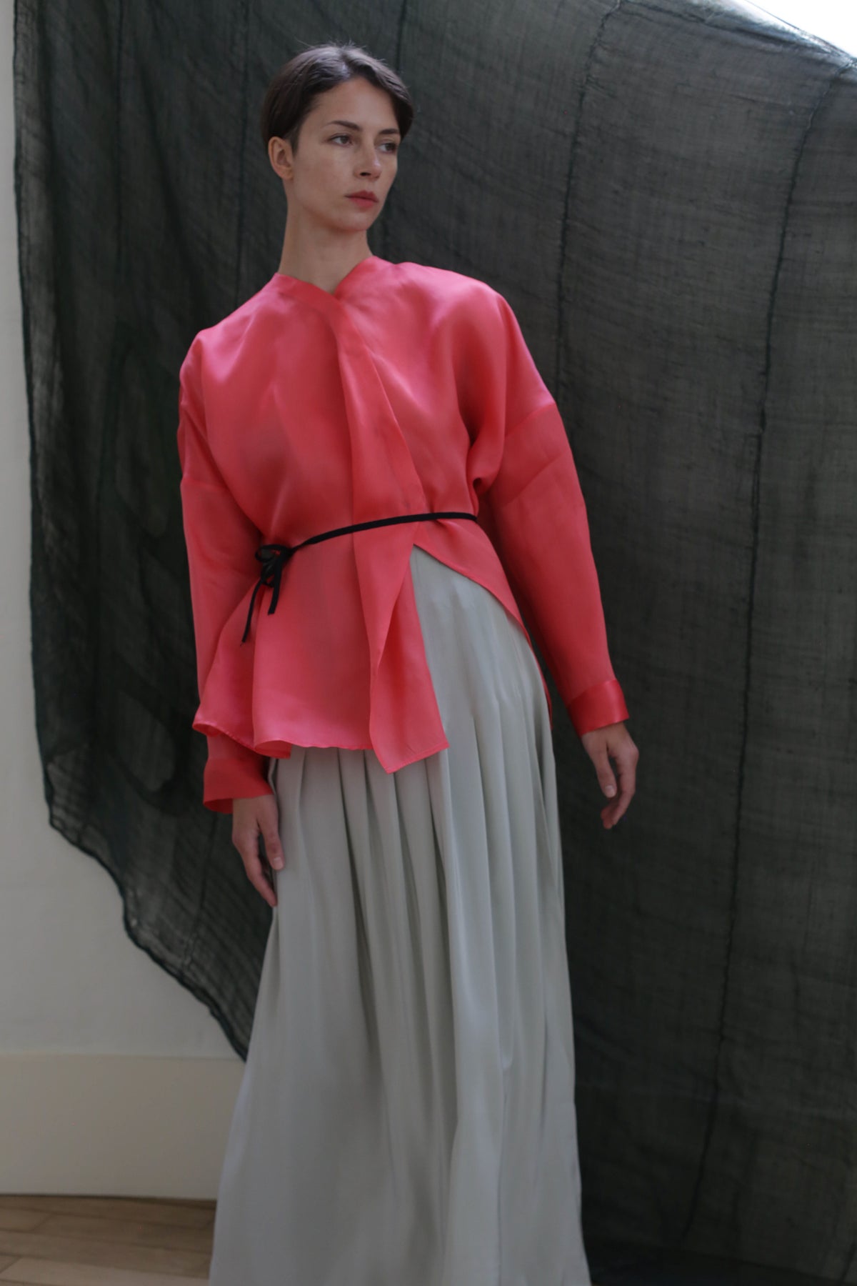 A person stands indoors wearing a loose, long-sleeve, bright pink blouse with a black belt and the Hand Pleated Column Wrap Skirt in Crepe de Chine Celadon. A sheer dark fabric hangs behind them in soft, natural lighting.