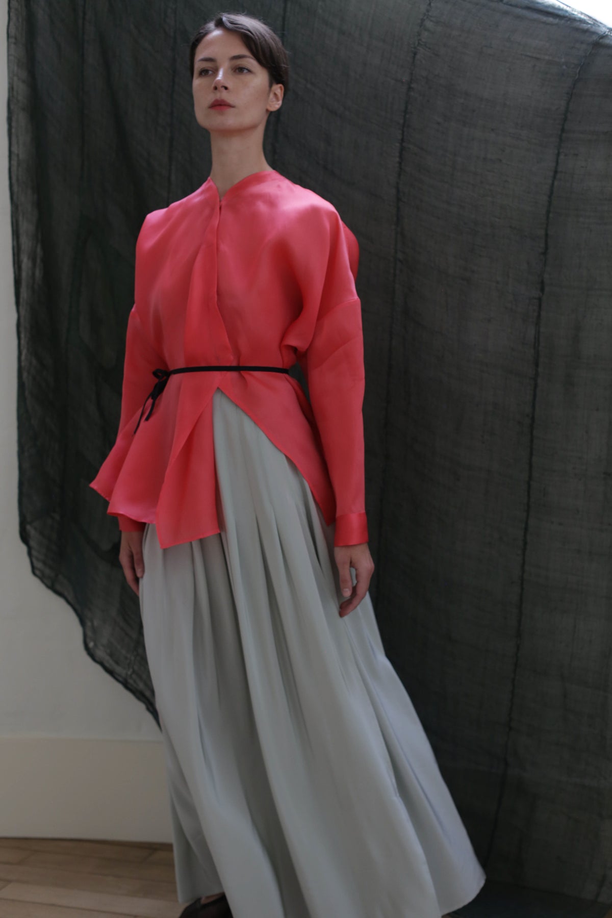 A person stands against a dark, sheer backdrop, dressed in a loose pink top with a black belt and the Hand Pleated Column Wrap Skirt in Crepe de Chine Celadon, featuring flowing lines inspired by zero-waste design.