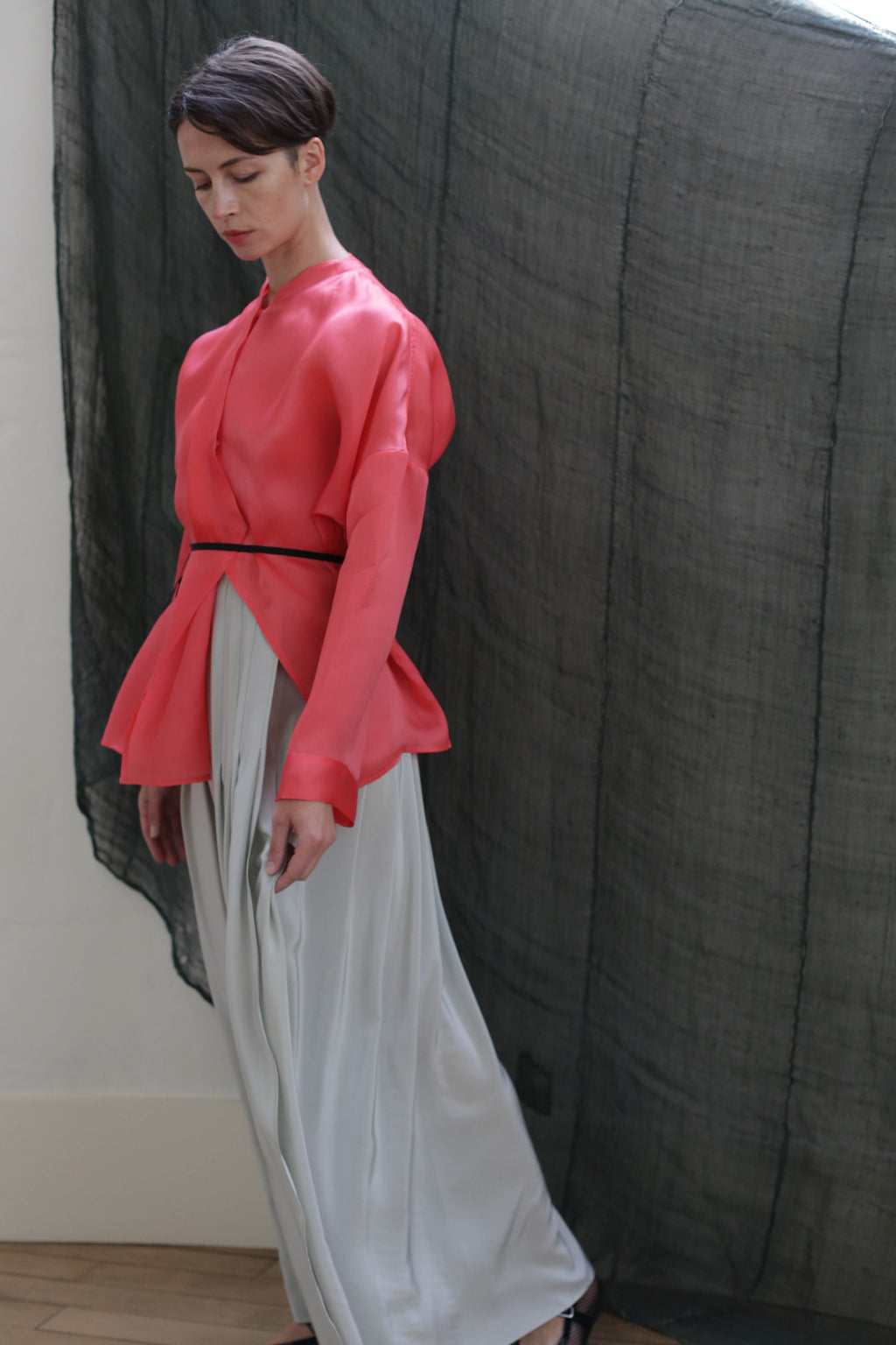 A person stands indoors in a coral pink long-sleeved top, black belt, and the Hand Pleated Column Wrap Skirt | Crepe de Chine Celadon, posed before a dark sheer fabric backdrop.