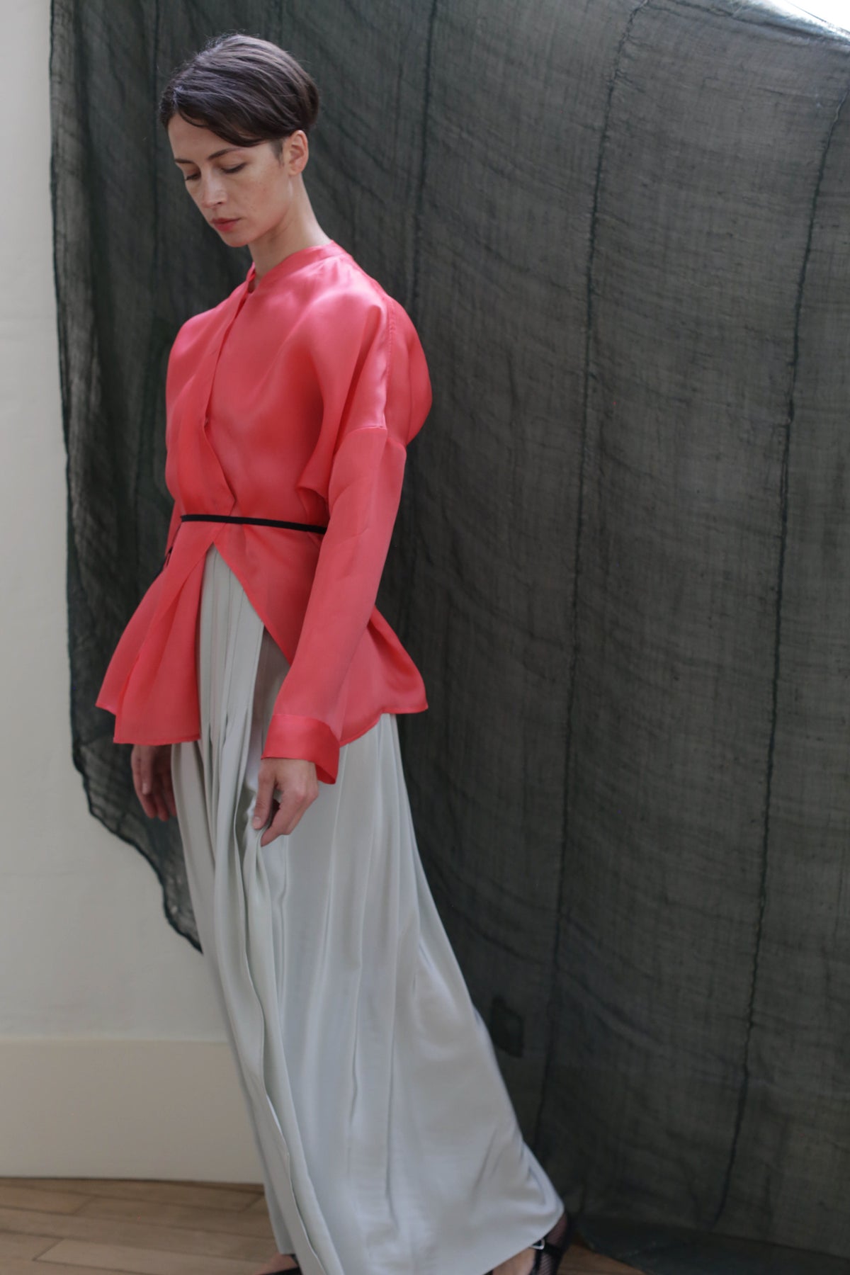A person stands indoors in a coral pink long-sleeved top, black belt, and the Hand Pleated Column Wrap Skirt | Crepe de Chine Celadon, posed before a dark sheer fabric backdrop.