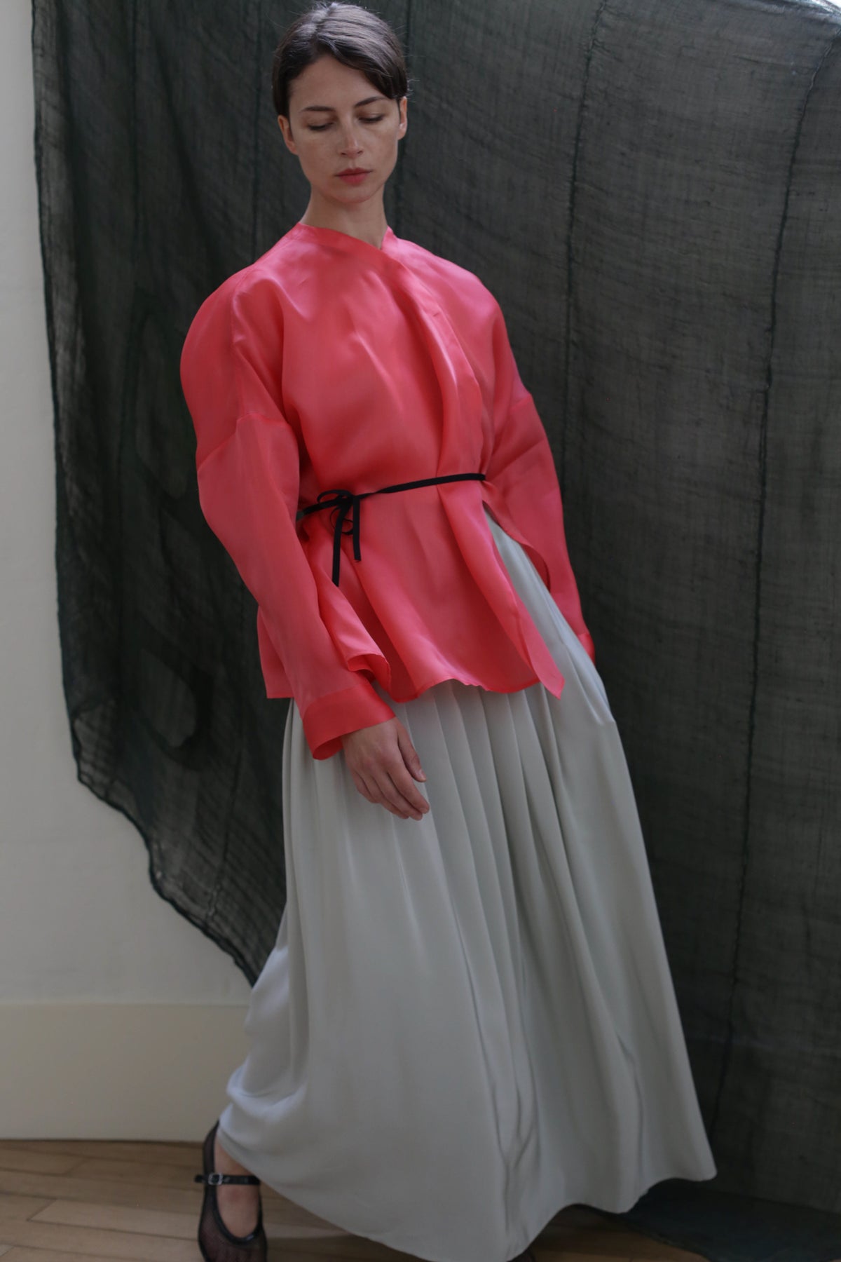 A person stands indoors before a dark, sheer backdrop, wearing a bright pink long-sleeved top with a black belt and the Hand Pleated Column Wrap Skirt in Crepe de Chine Celadon.