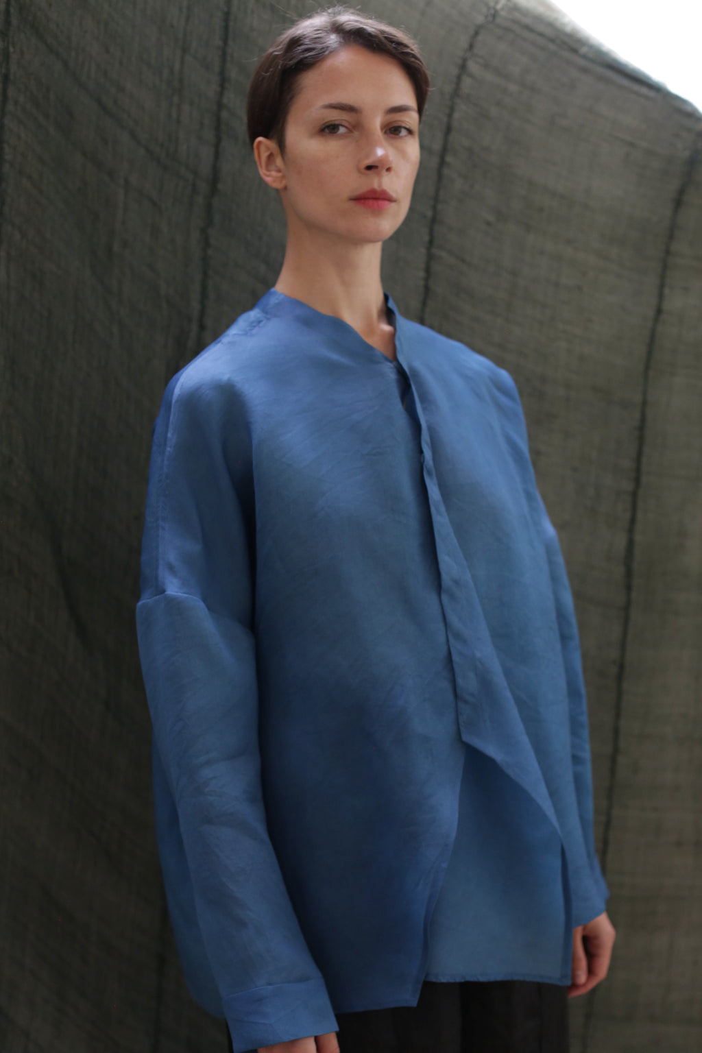 A woman with short brown hair wears the Qipao Blouse | Silk Organza Ai Dye—a loose-fitting, long-sleeved blue shirt—while standing before a textured dark green backdrop, facing slightly forward with a calm expression.
