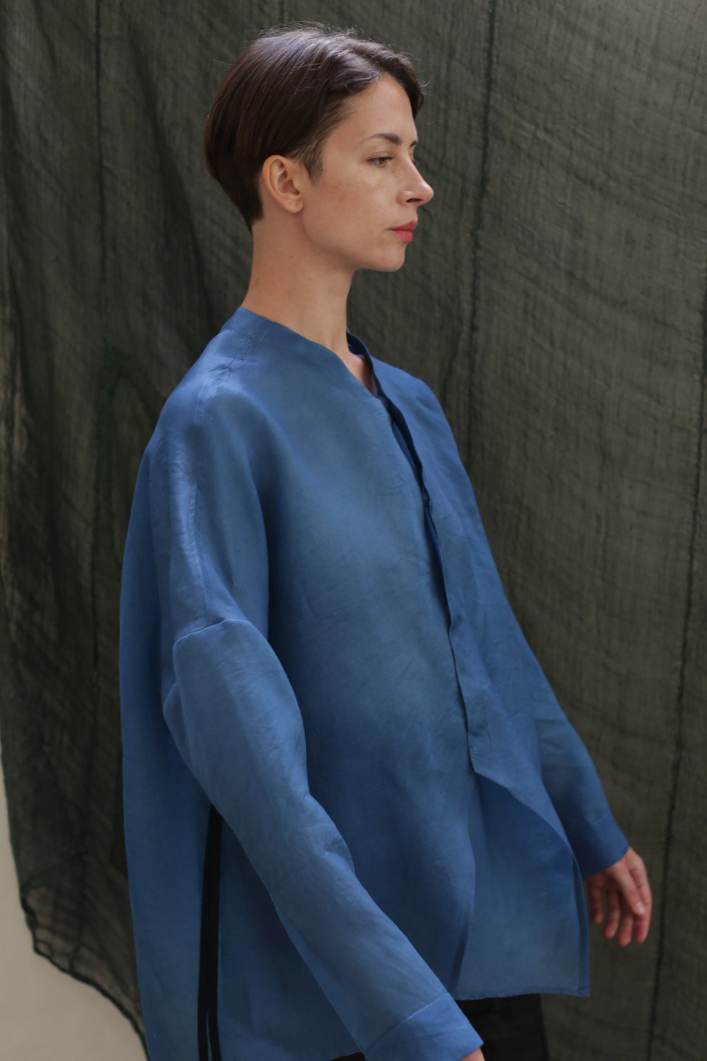 A person with short brown hair, wearing the Qipao Blouse in hand-dyed silk organza, stands in profile against a textured dark green fabric backdrop.