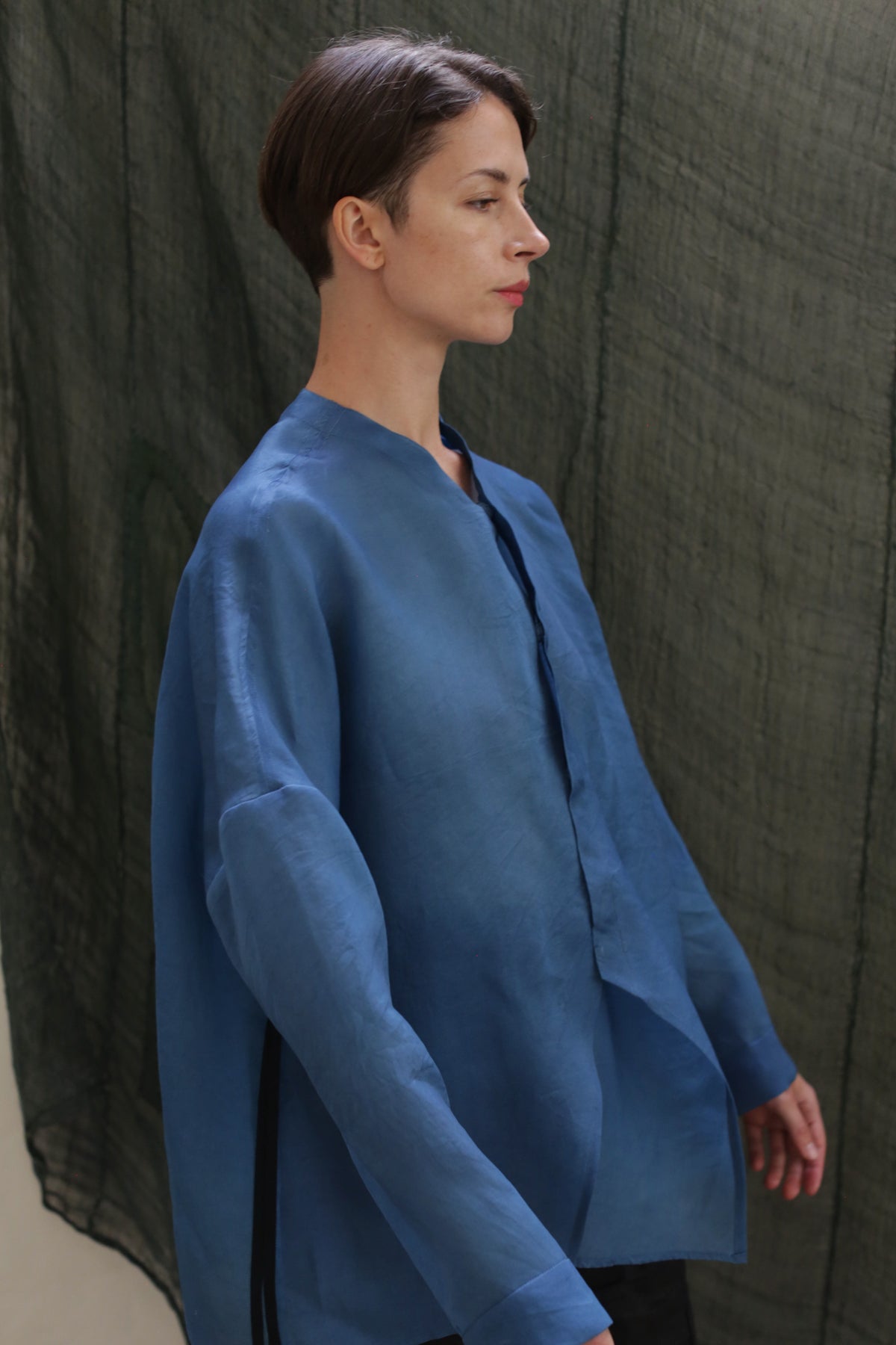 A person with short brown hair, wearing the Qipao Blouse in hand-dyed silk organza, stands in profile against a textured dark green fabric backdrop.