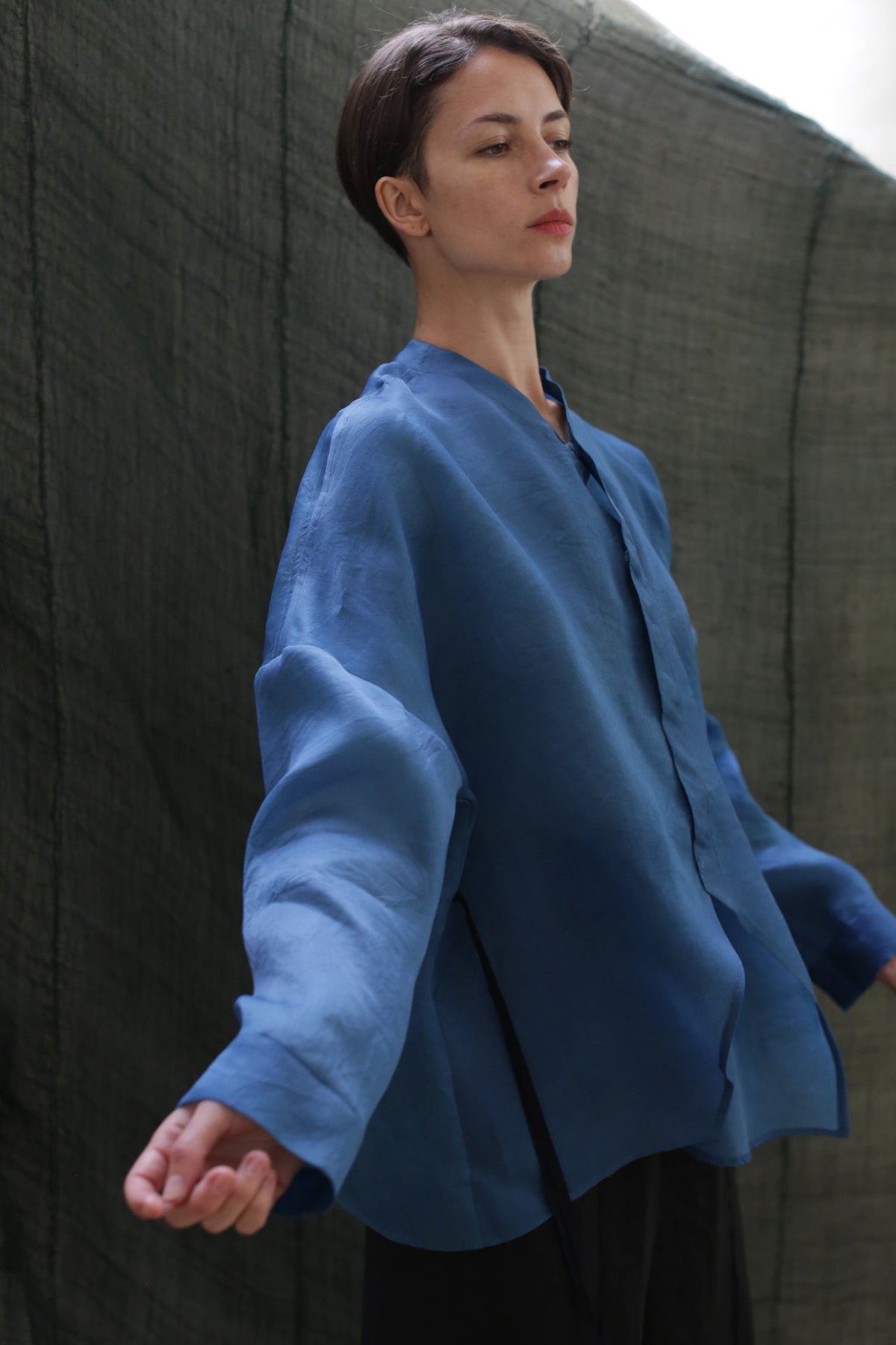 A person with short hair poses in front of a dark, textured backdrop wearing the Qipao Blouse | Silk Organza Ai Dye—an oversized, blue hand-dyed button-up with wide sleeves, arms slightly extended and head gently tilted back.