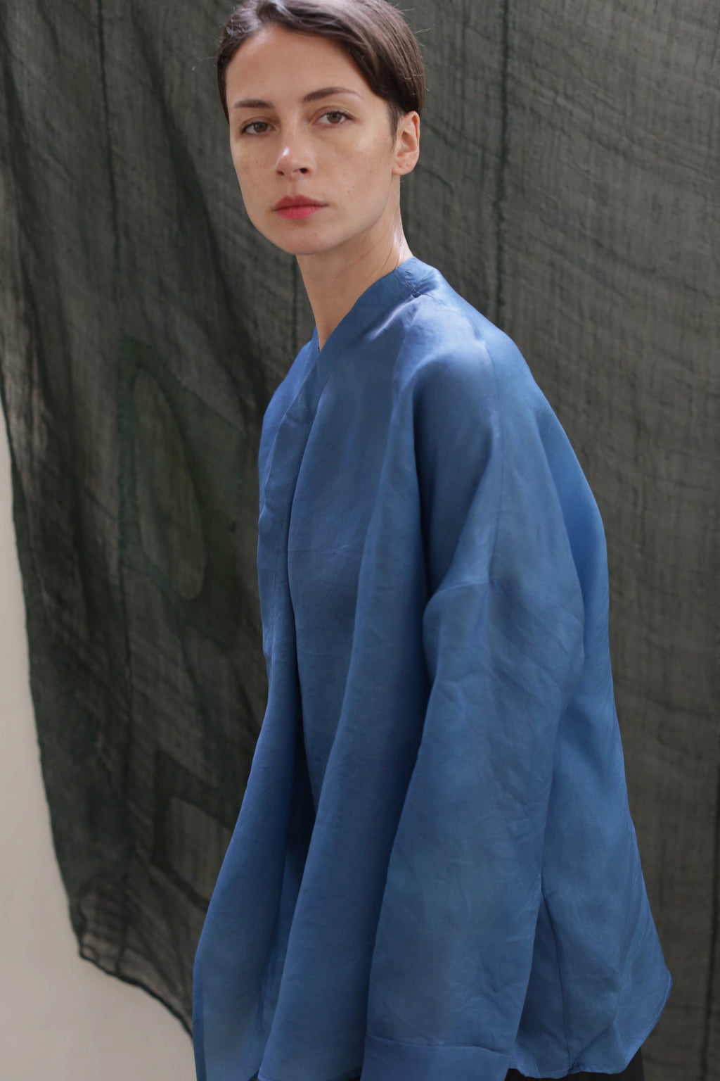 A person with short brown hair and red lipstick wears the Qipao Blouse in hand-dyed blue silk organza, posing before a dark green textured fabric backdrop.
