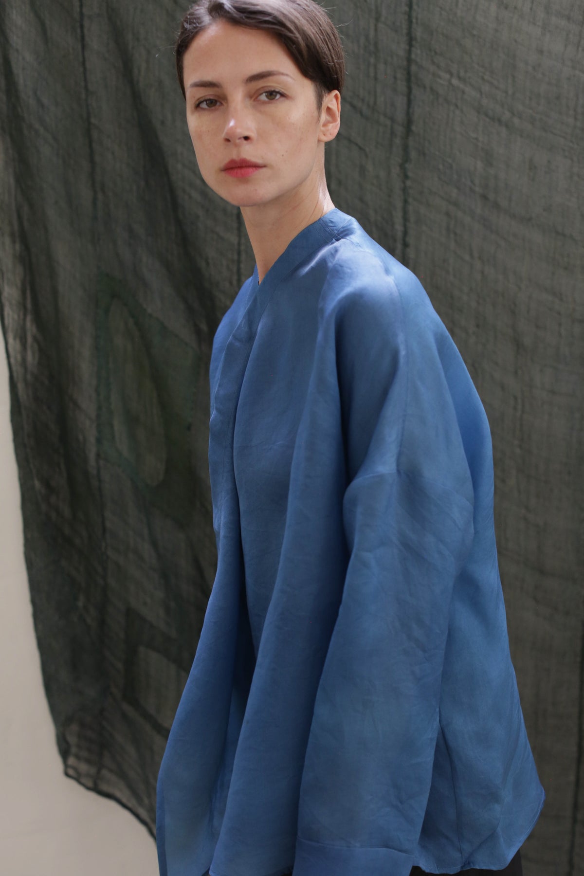 A person with short brown hair and red lipstick wears the Qipao Blouse in hand-dyed blue silk organza, posing before a dark green textured fabric backdrop.
