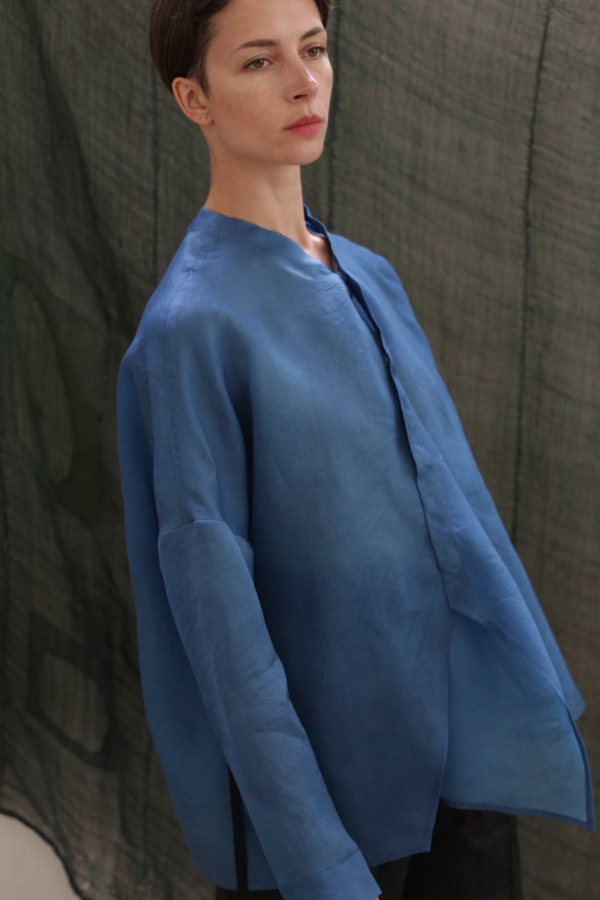A person with short brown hair wears the Qipao Blouse | Silk Organza Ai Dye, a loose-fitting, long-sleeved hand-dyed blue shirt, standing before a textured dark green backdrop and looking slightly to the side with a neutral expression.