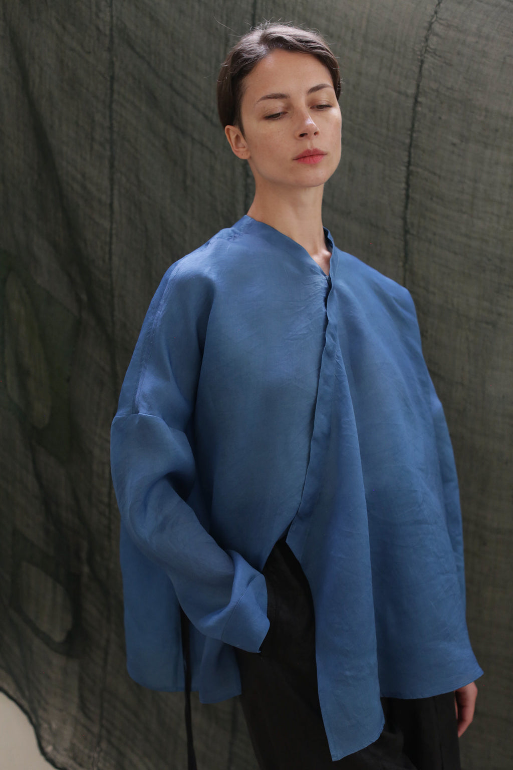 A woman with short brown hair, wearing the oversized Qipao Blouse | Silk Organza Ai Dye in blue and black pants, stands with one hand in her pocket against draped green fabric, her eyes closed in a calm, relaxed pose.