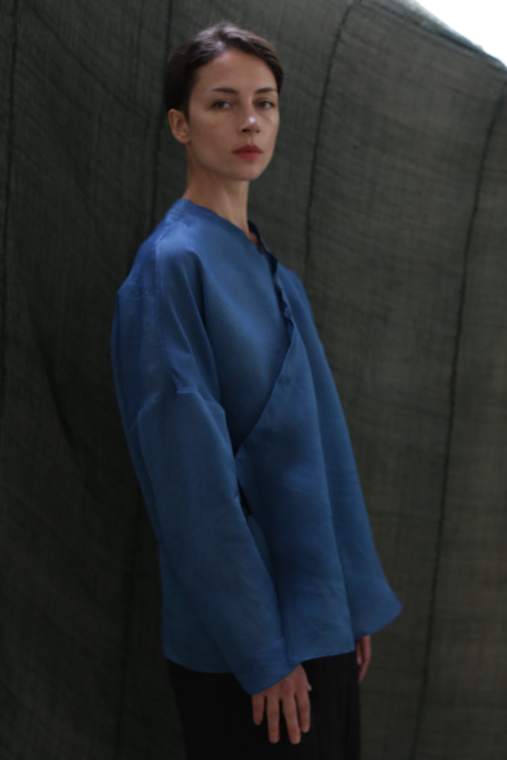 A woman with short dark hair stands against a dark green fabric backdrop, wearing the Qipao Blouse in hand-dyed silk organza and gazing calmly to the side.