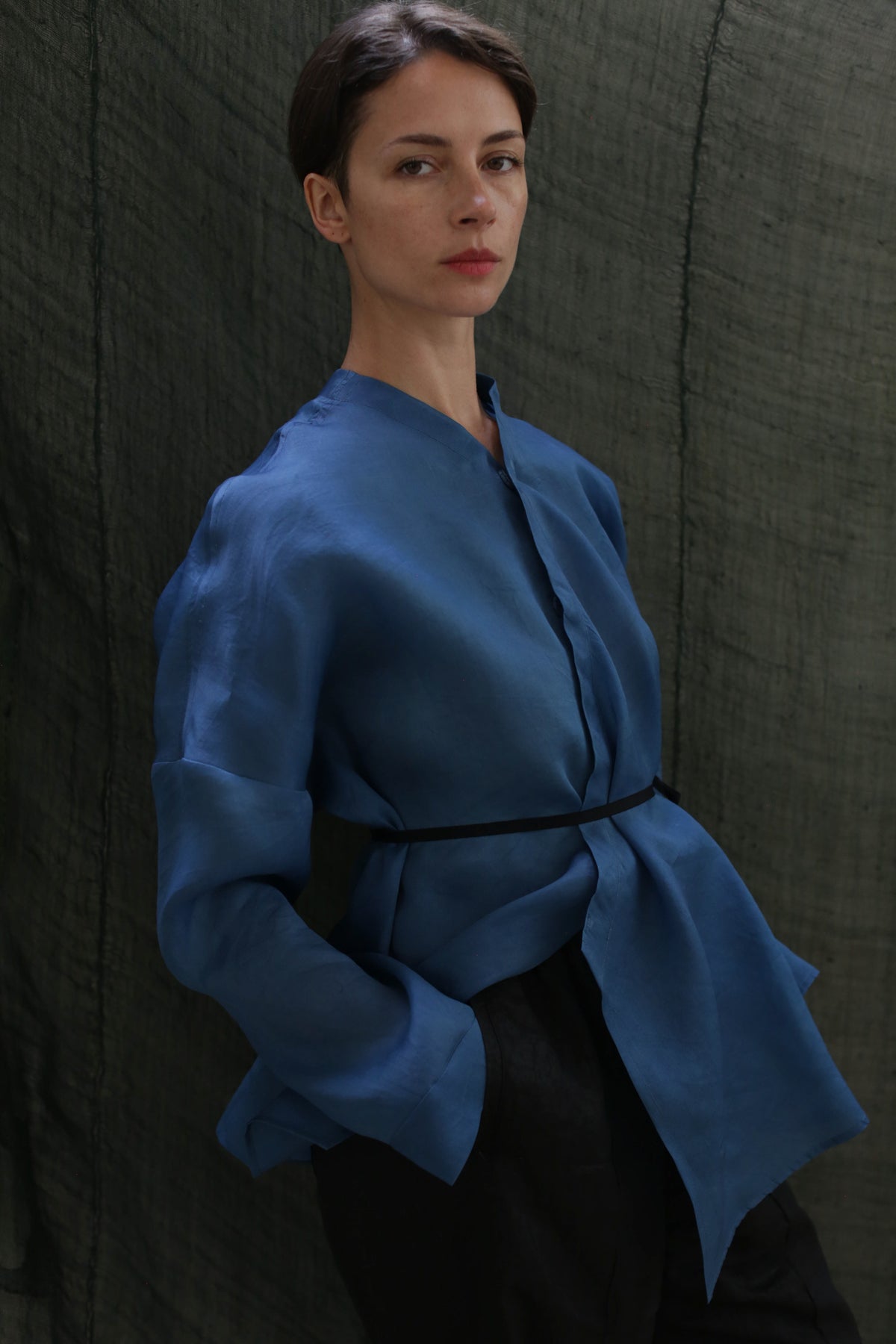 A woman with short dark hair wears the Qipao Blouse in hand-dyed blue silk organza, cinched at the waist with a thin black belt, standing against a dark green textured fabric backdrop.