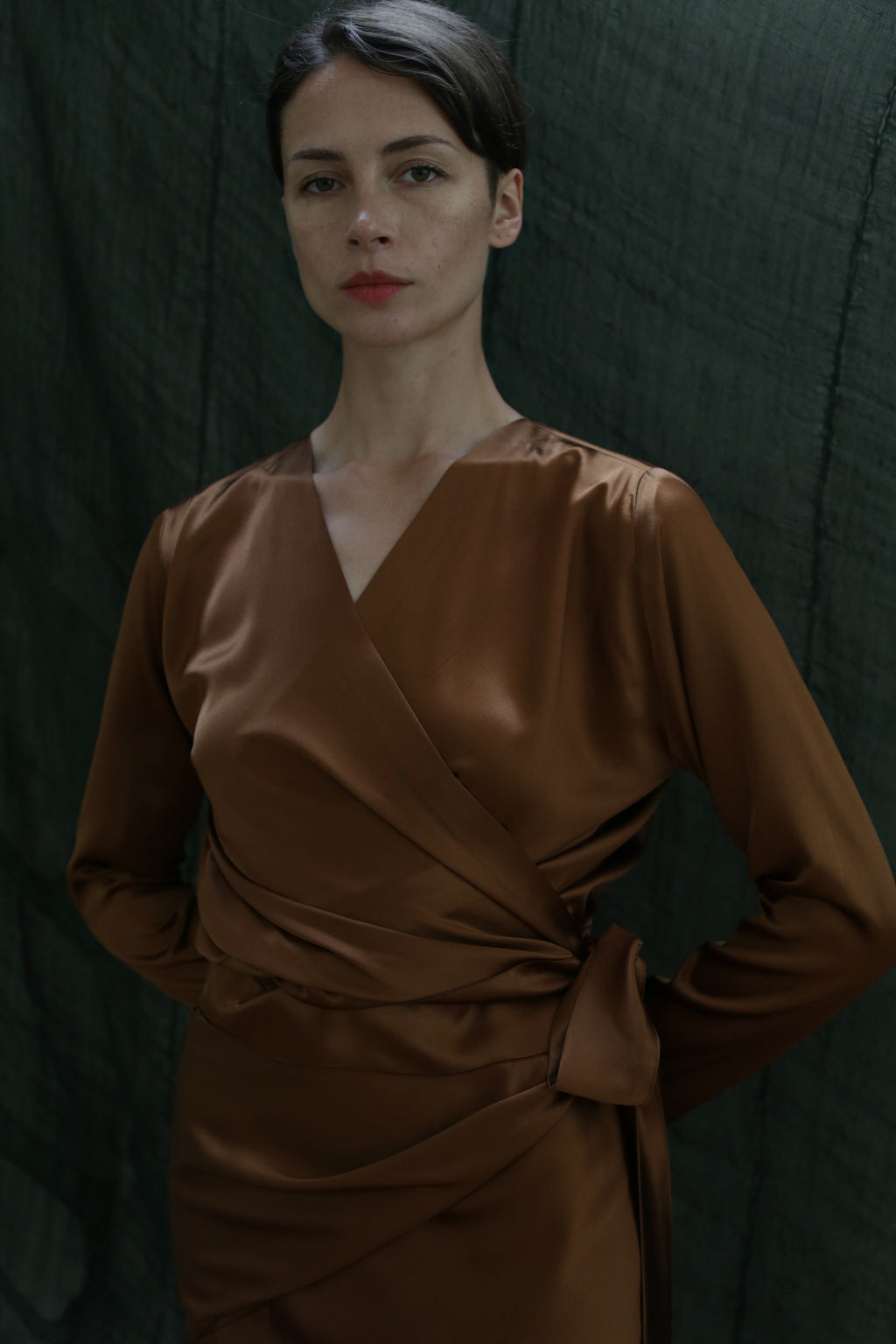 A woman with dark hair tied back stands before a dark green fabric backdrop, wearing the X Blouse in Silk Charmeuse Copper, long-sleeved with a sash at the waist, calmly looking at the camera.
