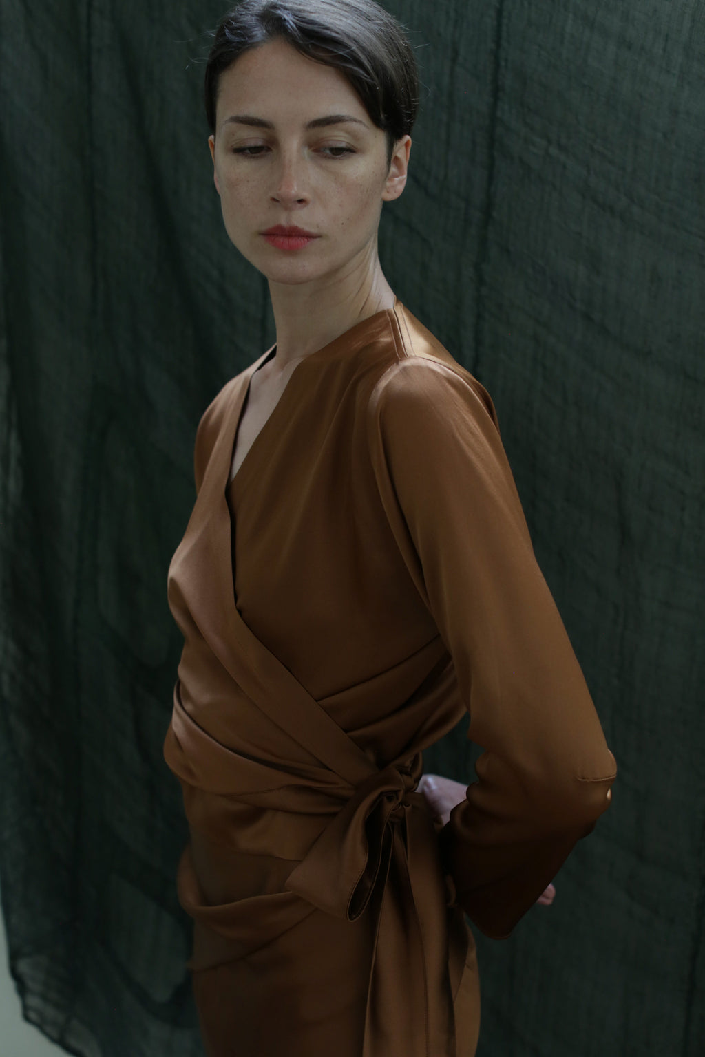 A woman wears the X Blouse in Silk Charmeuse Copper, standing before a dark green textured backdrop, looking slightly down and to the side with a calm, neutral expression.