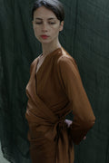 A woman wears the X Blouse in Silk Charmeuse Copper, standing before a dark green textured backdrop, looking slightly down and to the side with a calm, neutral expression.