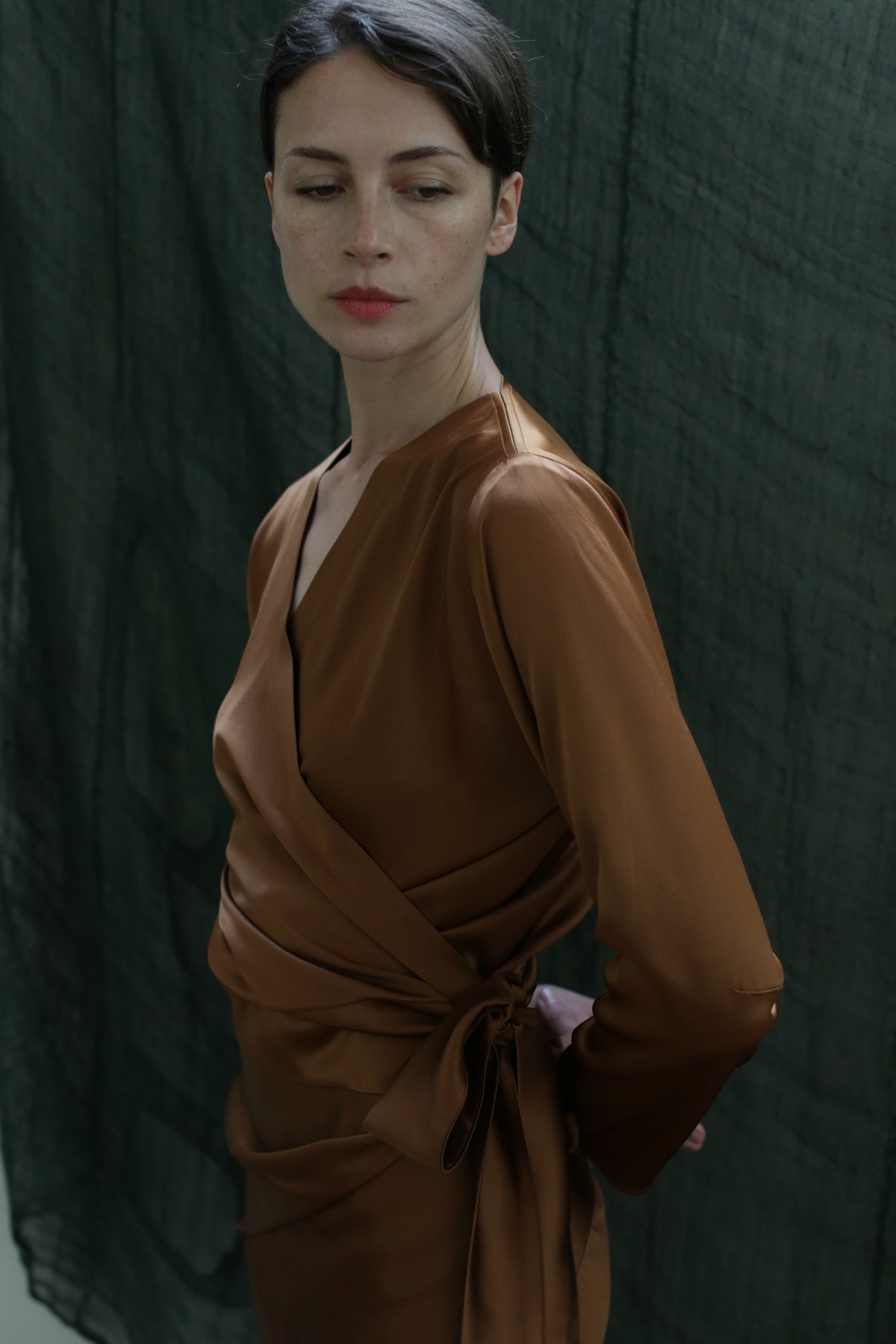 A woman wears the X Blouse in Silk Charmeuse Copper, standing before a dark green textured backdrop, looking slightly down and to the side with a calm, neutral expression.