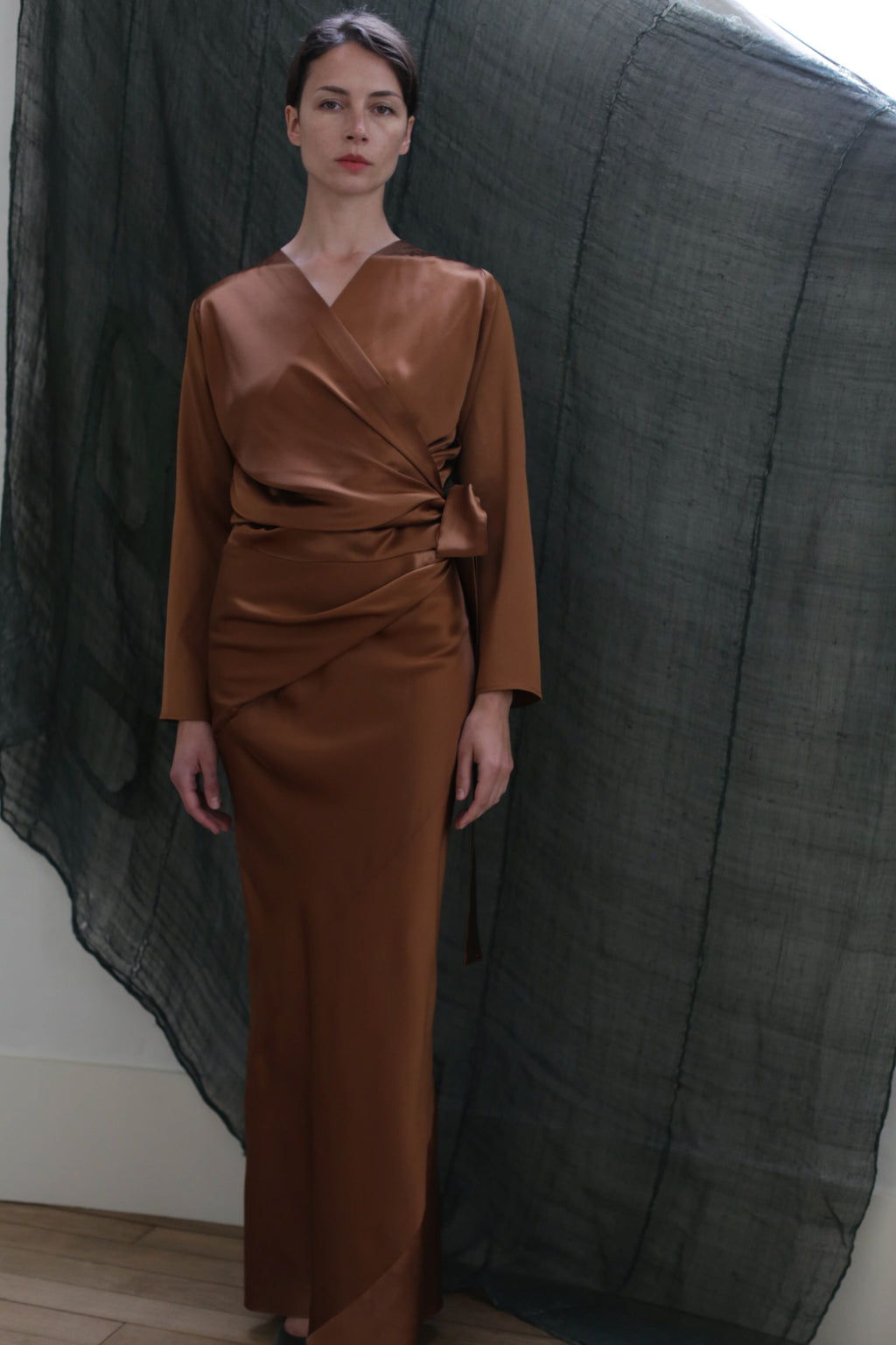 A woman stands before a draped dark backdrop, wearing Atelier Lan Jaenicke's Dragon Skirt in Silk Charmeuse Copper, featuring a side tie and floor-length silhouette.