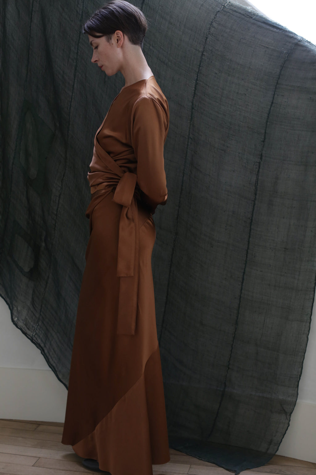 A woman with short dark hair stands in profile, wearing the X Blouse in Silk Charmeuse Copper, featuring a tied waist and elegant drape, against a sheer dark green fabric backdrop.